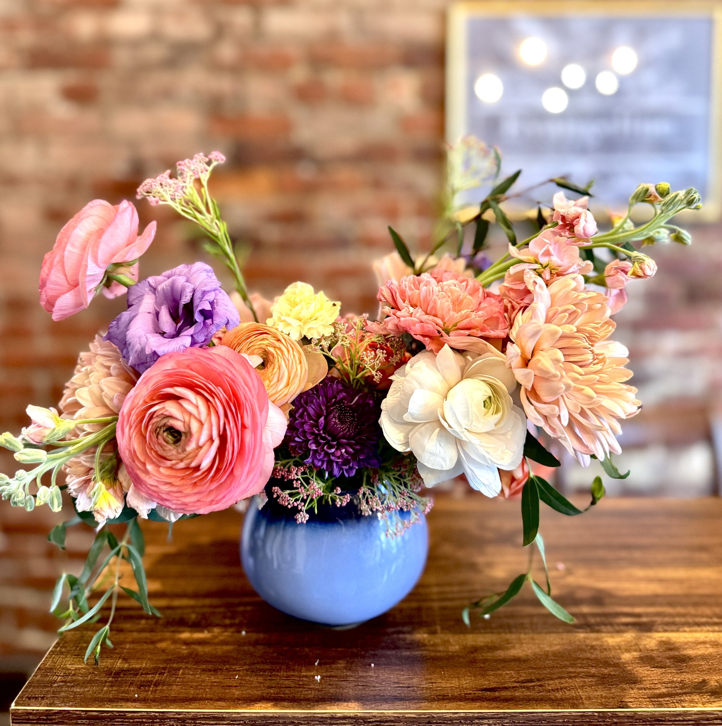 Colorful floral arrangement in a blue vase on a wooden table with a brick wall background.