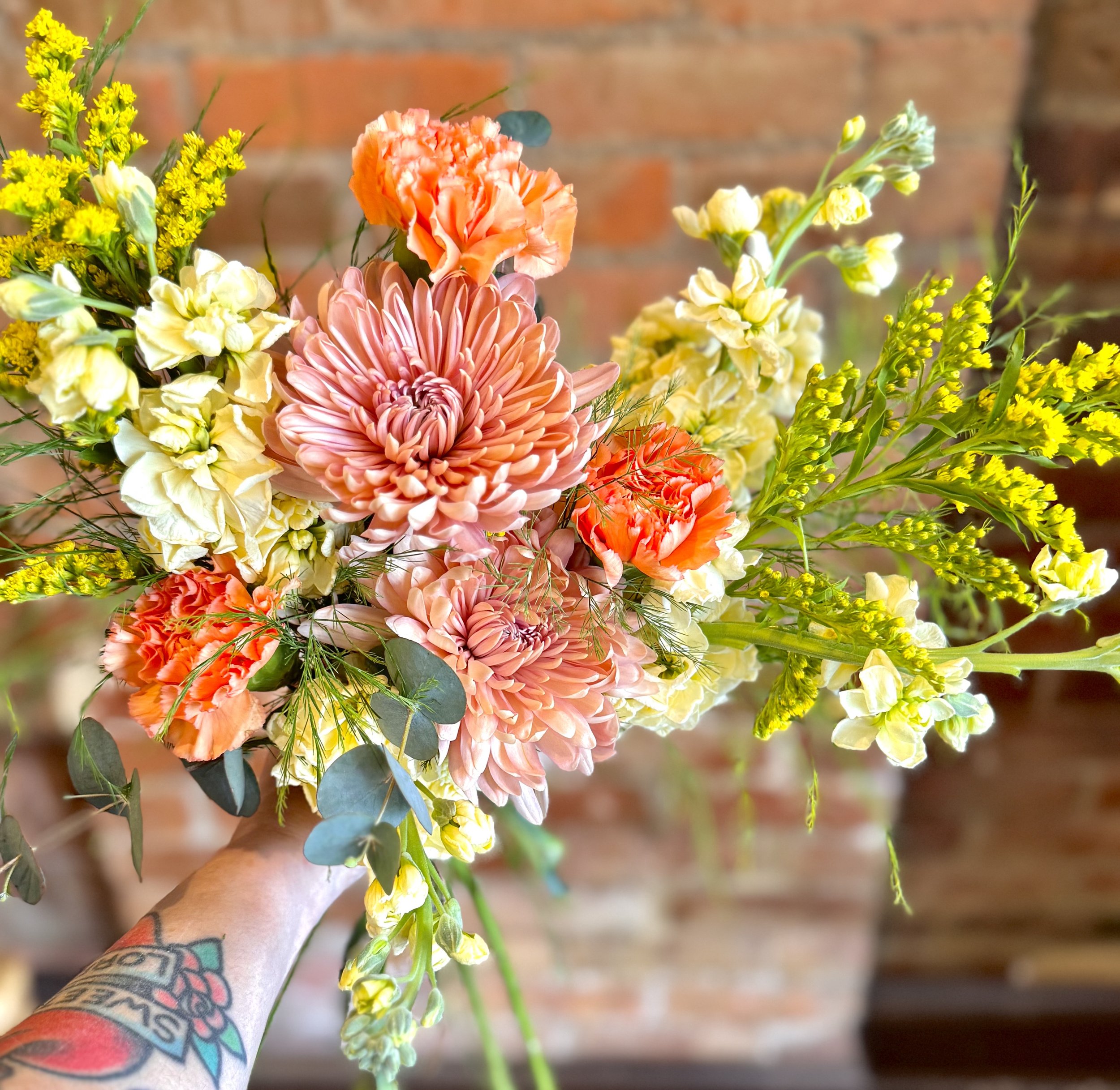 Bouquet with light pink chrysanthemums, orange carnations, yellow flowers, and greenery against brick background.