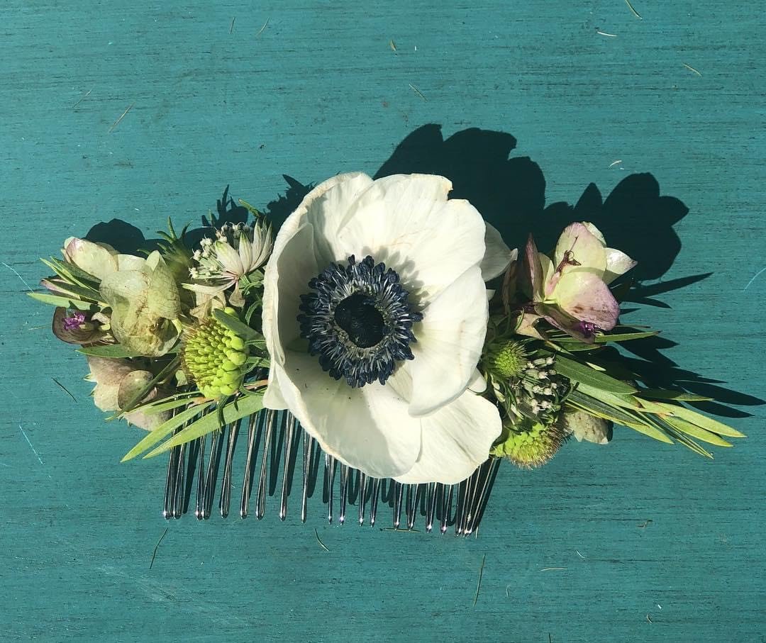 Floral hair comb with white anemone and green leaves on teal surface.