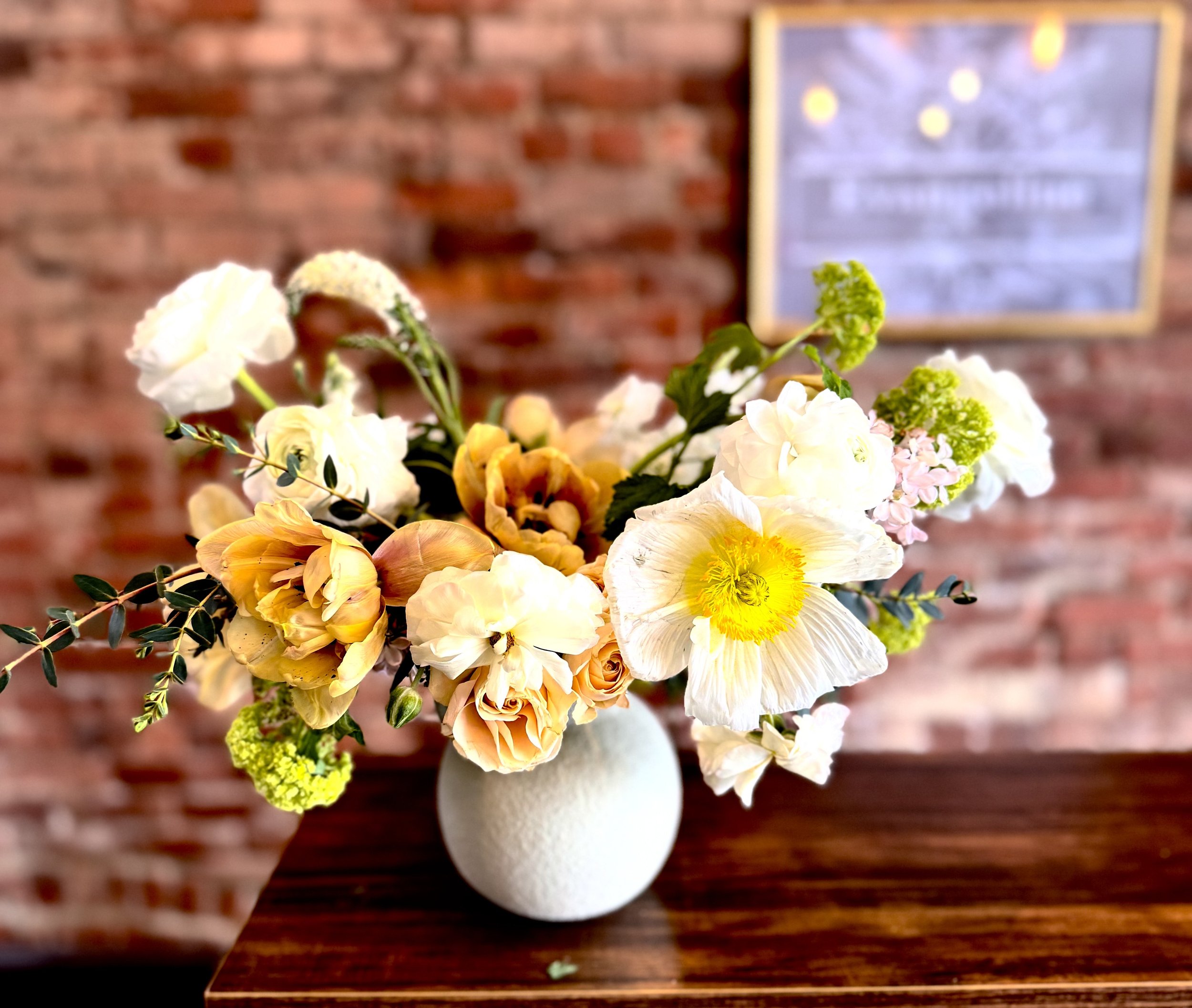 White ceramic vase with a mixed flower arrangement, featuring white and yellow blooms, on a wooden table against a brick wall.