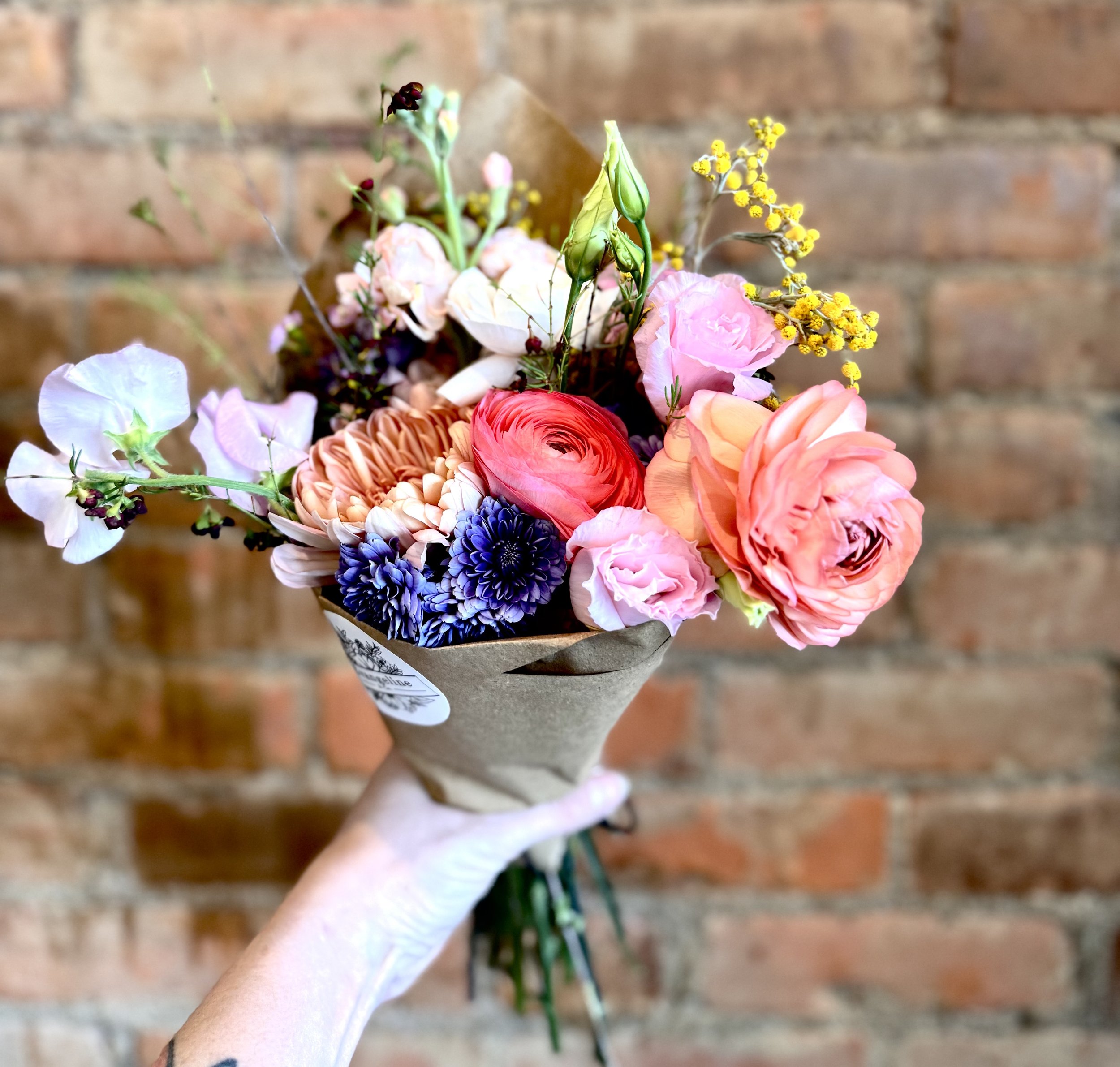 Colorful bouquet of assorted flowers, including pink and orange roses, wrapped in brown paper, against a brick wall background.