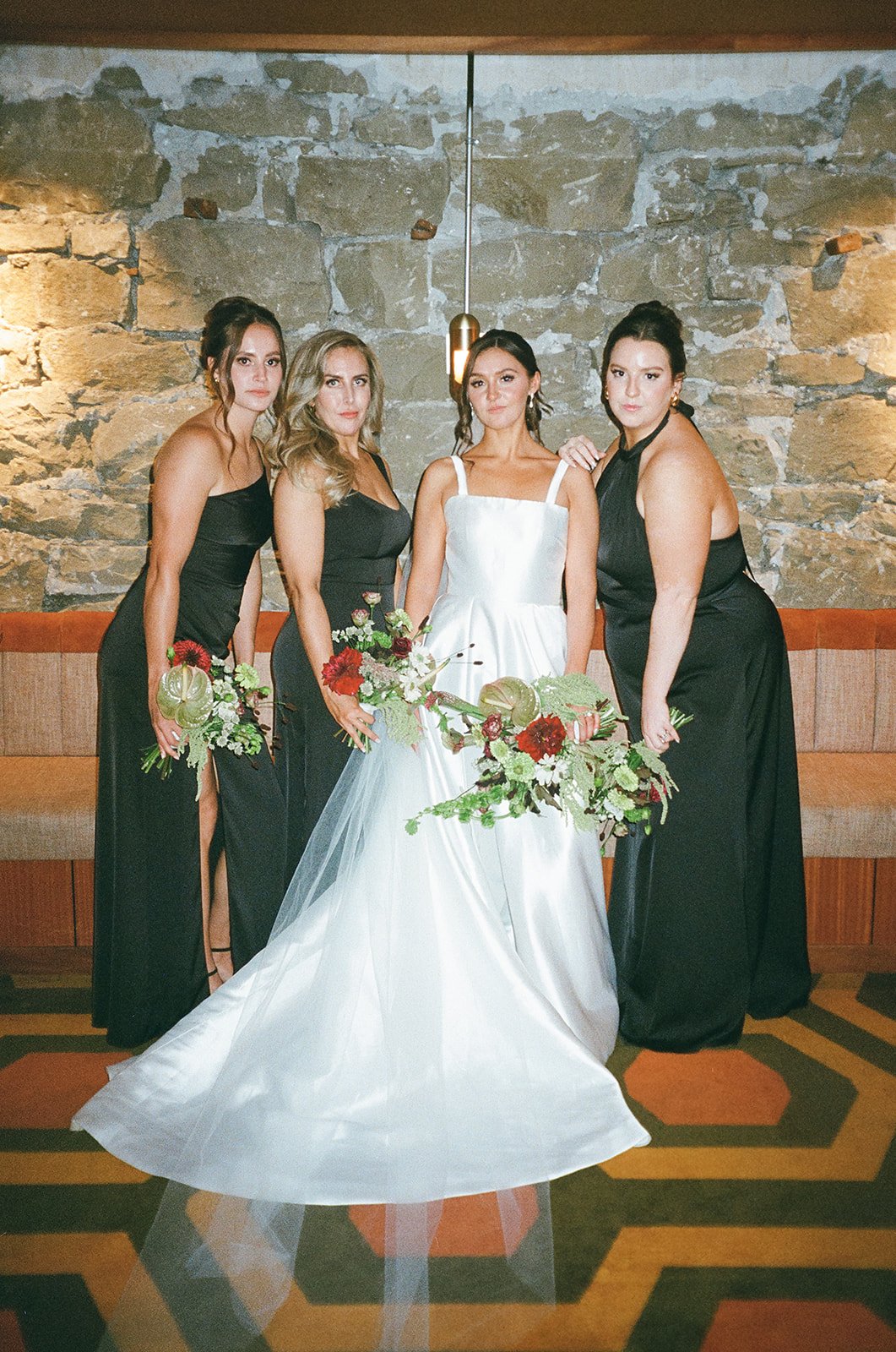Bride and bridesmaids posing indoors with stone wall background, bride in white gown holding bouquet, bridesmaids in black dresses holding flowers.
