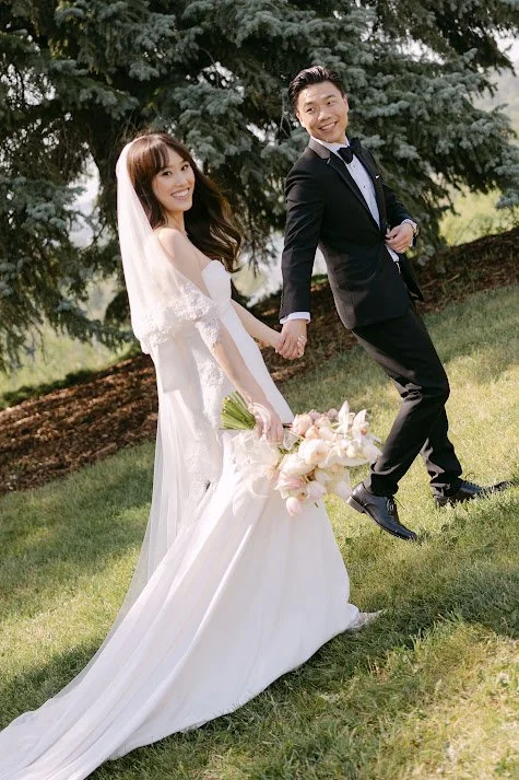 A bride and groom walking hand in hand outdoors, both smiling. The bride is wearing a long white wedding dress with a veil and holding a bouquet of flowers. The groom is dressed in a black tuxedo. They are on grass with a tree in the background.