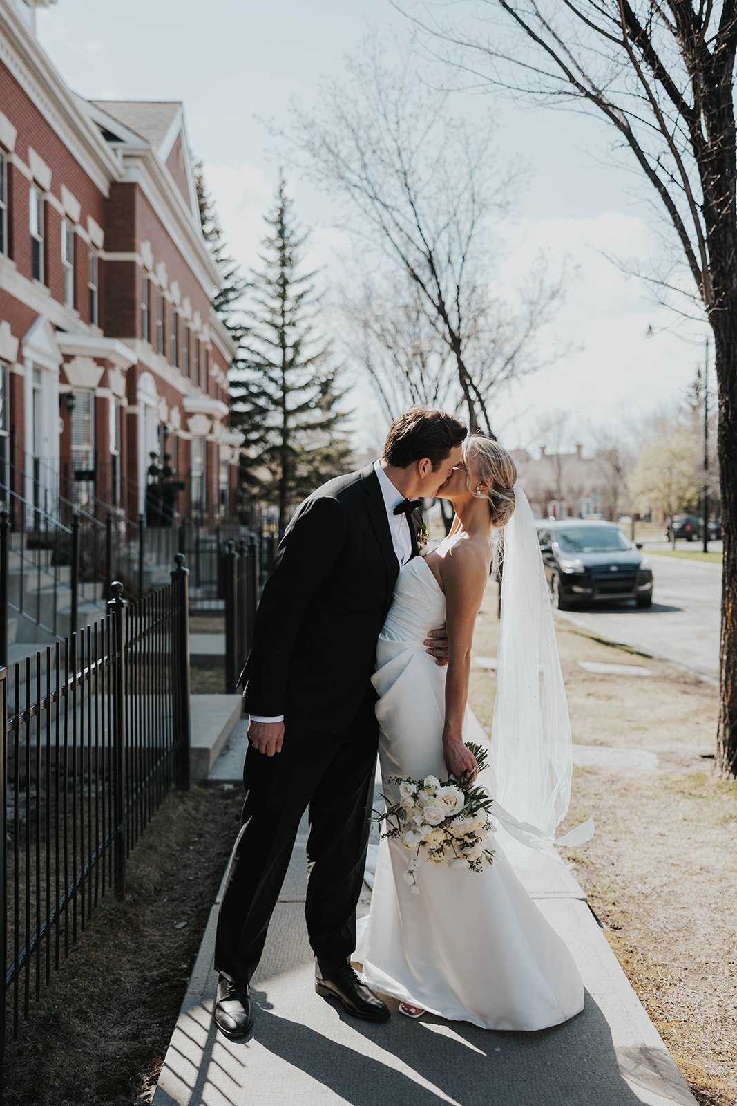 Bride and groom kissing on a sidewalk, bride holding bouquet, wearing wedding attire, urban setting with trees and buildings in the background.