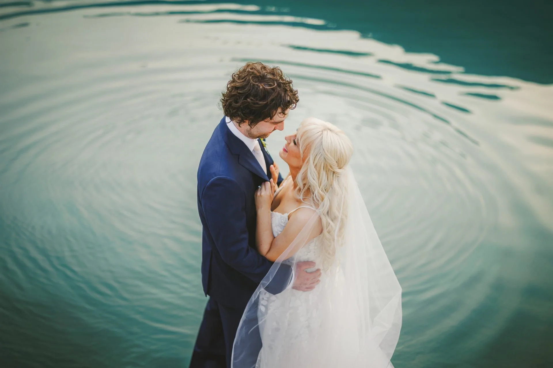 Bride and groom embracing near a body of water, with rippling water creating a serene backdrop; bride wearing a white dress and veil, groom in a dark suit.