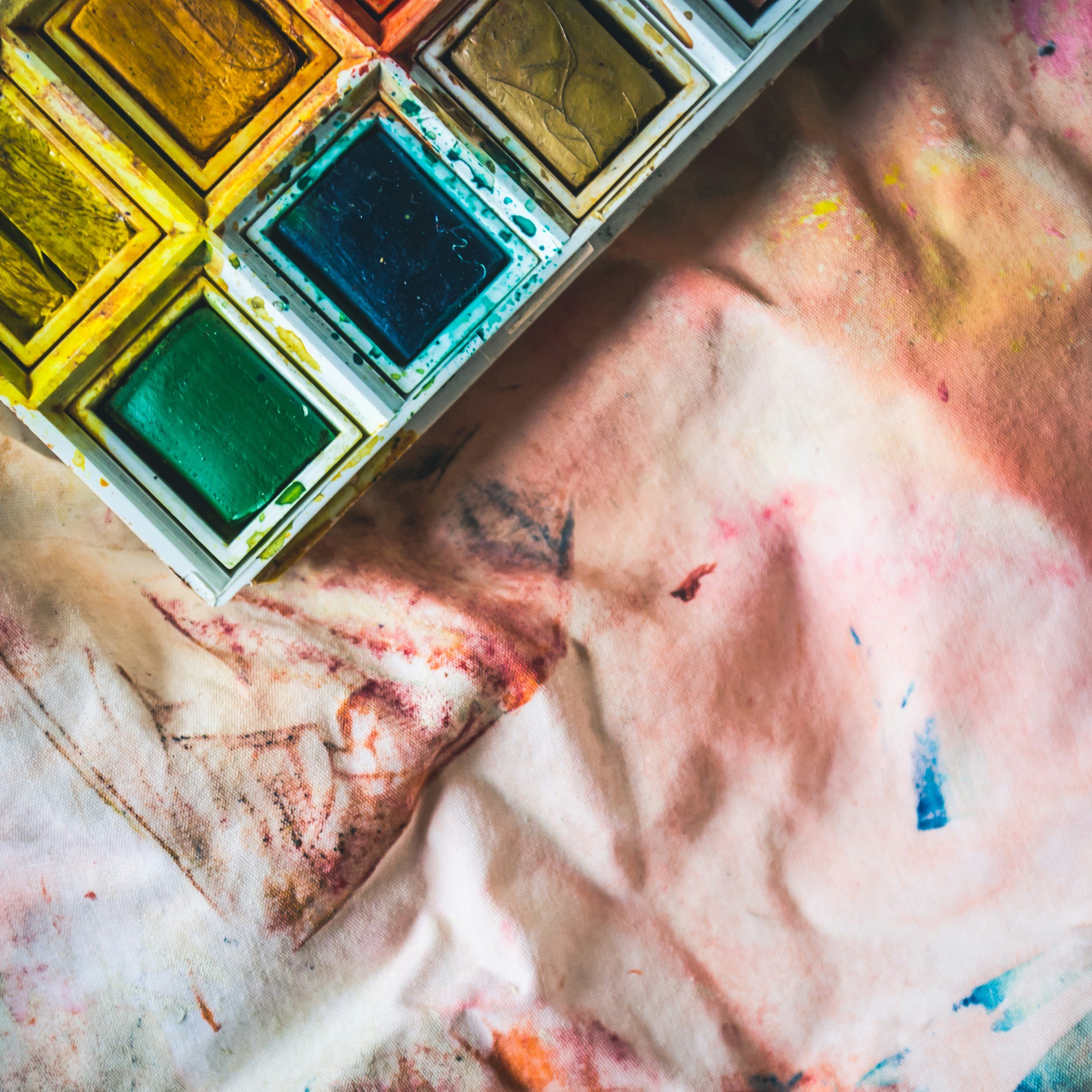 A watercolor paint palette with various colors, including yellow, green, blue, and brown, next to a cloth covered in multicolored paint stains.