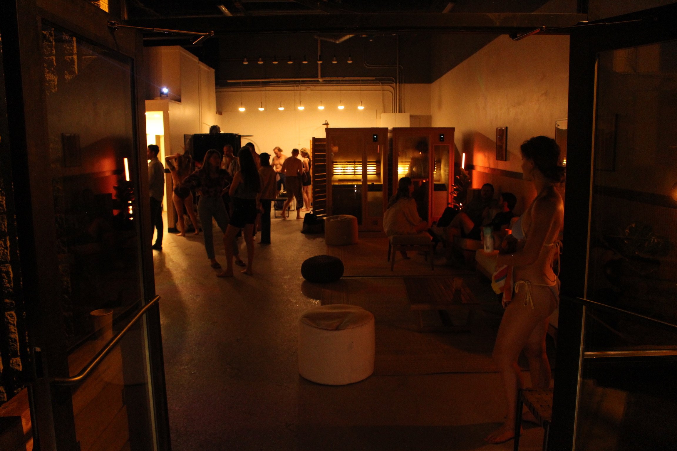 People gathering and socializing in a dimly lit indoor lounge or party space with warm lighting, seating areas, and a partition in the background.