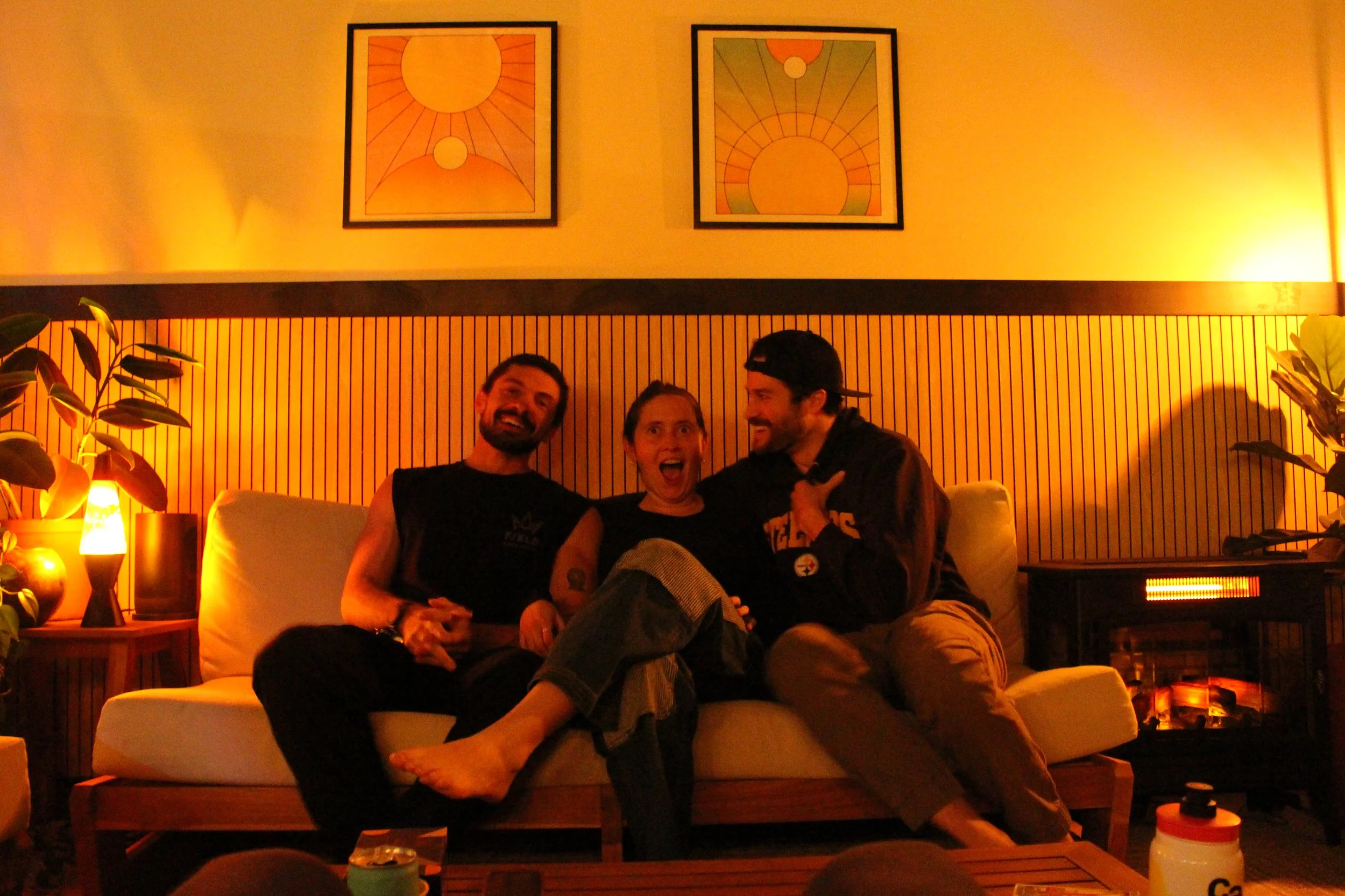 Three people sitting on a couch, smiling and laughing. The room is warmly lit, with two framed abstract art pieces hanging on the wall behind them and houseplants nearby.