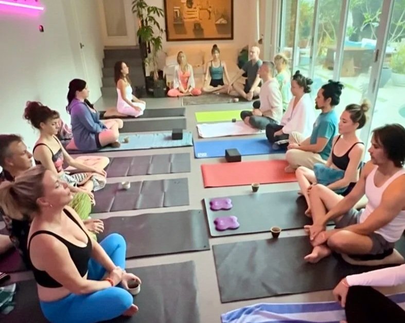 Group of people practicing yoga on mats in a bright indoor space with large windows and green plants.
