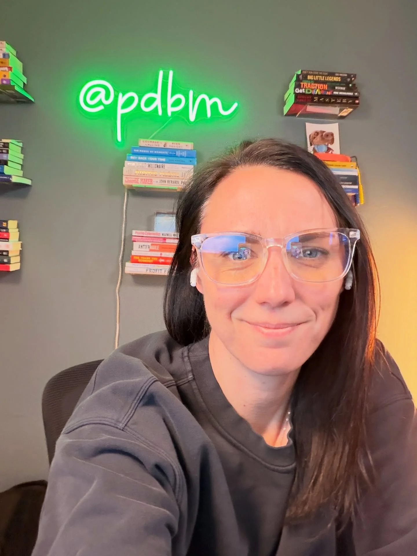 white woman with brown hair and glasses sits in front of a green wall with books and a green neon sign that says "@pdbm"