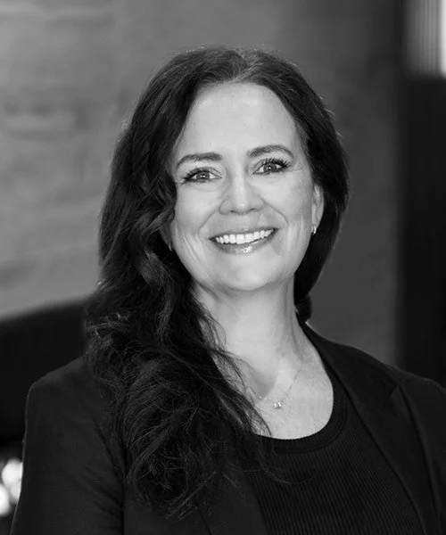 Black and white portrait of a smiling woman with long wavy hair, wearing a dark top and a necklace.