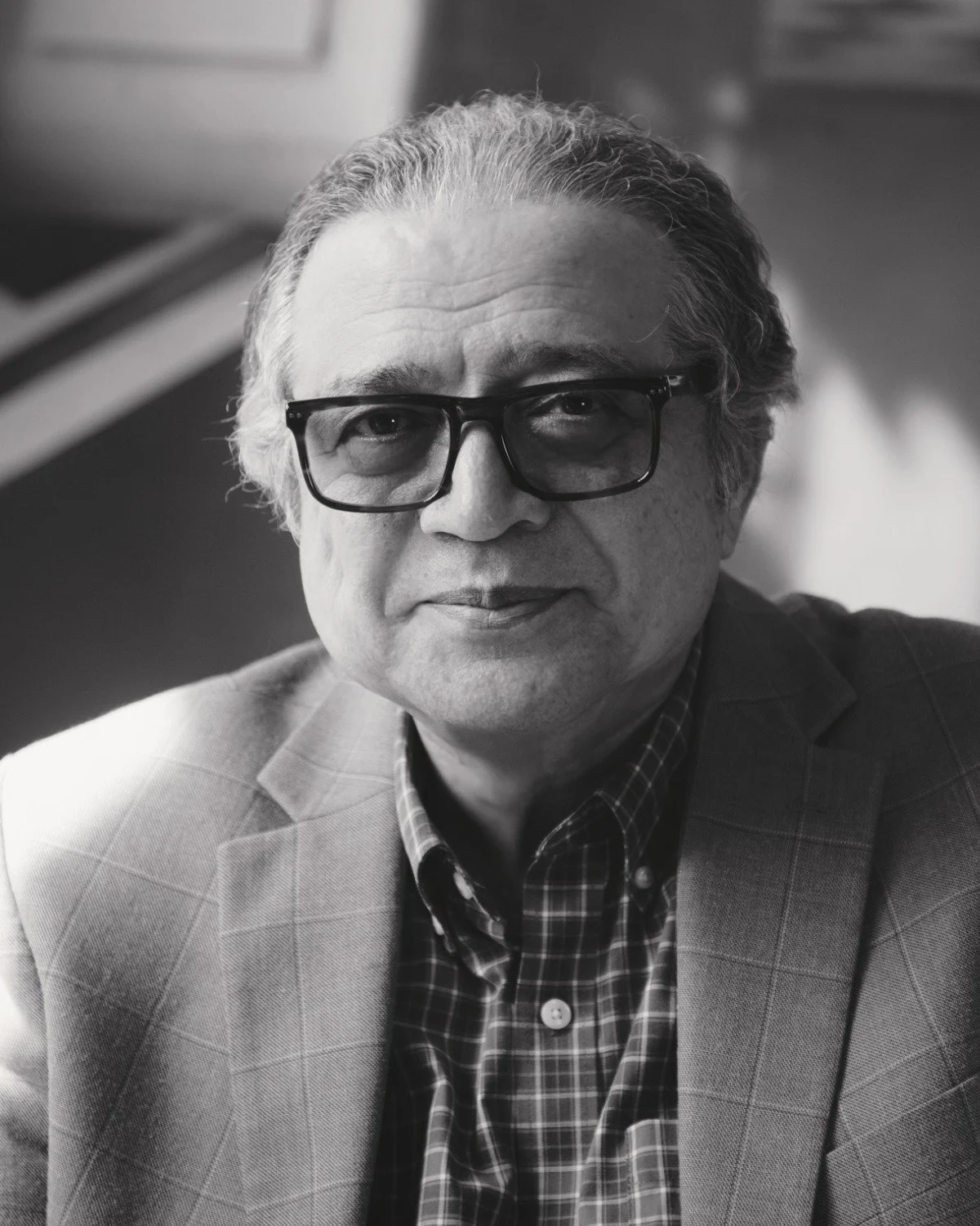 A black-and-white portrait of a middle-aged man wearing glasses, a checked shirt, and a suit jacket, smiling slightly.