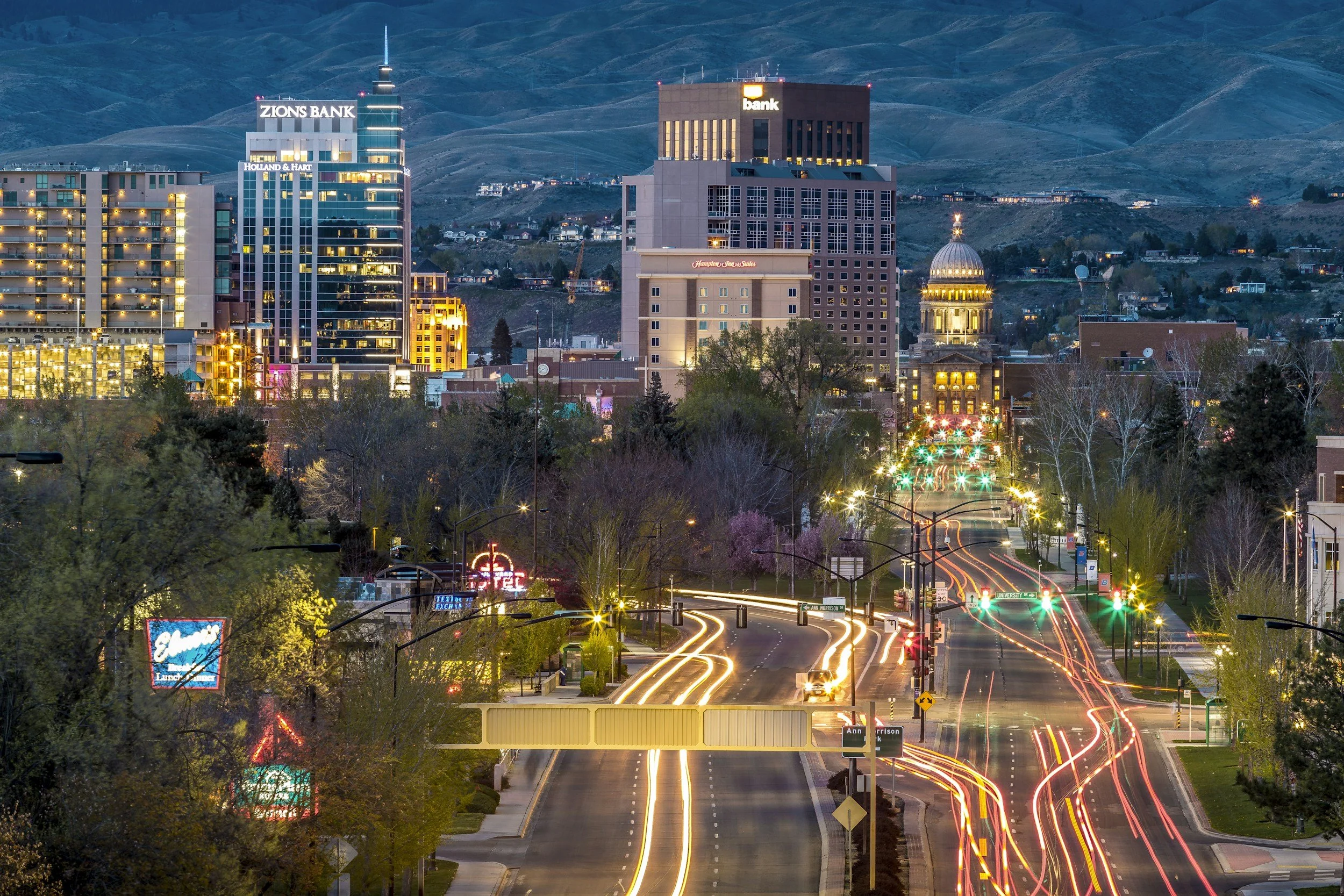 Night view of Boise, Idaho skyline featuring Zions Bank and other buildings, with streaks of car lights on a street lined with trees and neon signs, against a backdrop of hills.