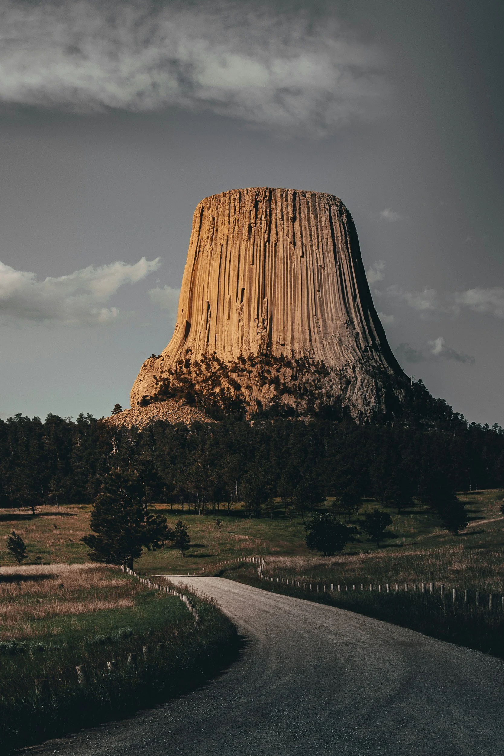 Devils Tower National Monument with road and forest, under cloudy sky.