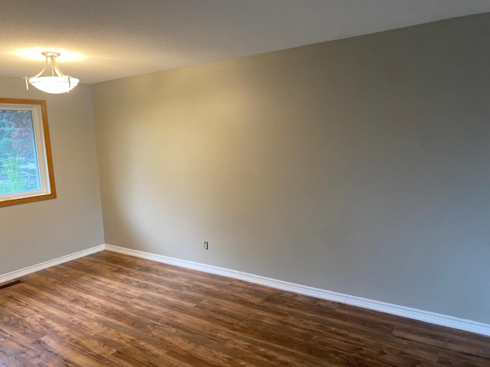Empty room with a wooden floor, a window with wood trim, and a ceiling light fixture.