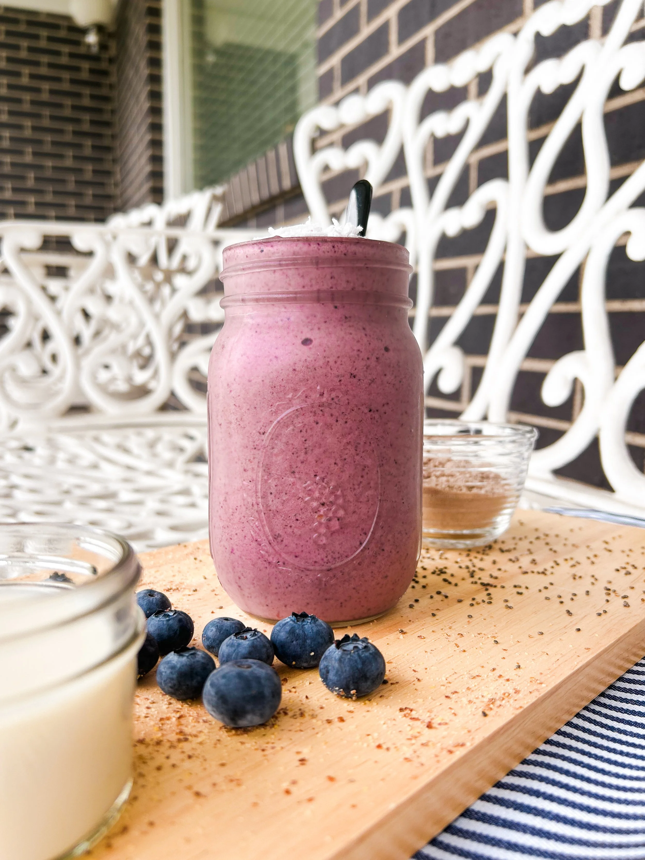 A fruit smoothie in a mason jar glass with a spoon inside, placed on a wooden board with fresh blueberries and small bowls of milk and protein powder against a white decorative metal bench and an outdoor brick wall background.
