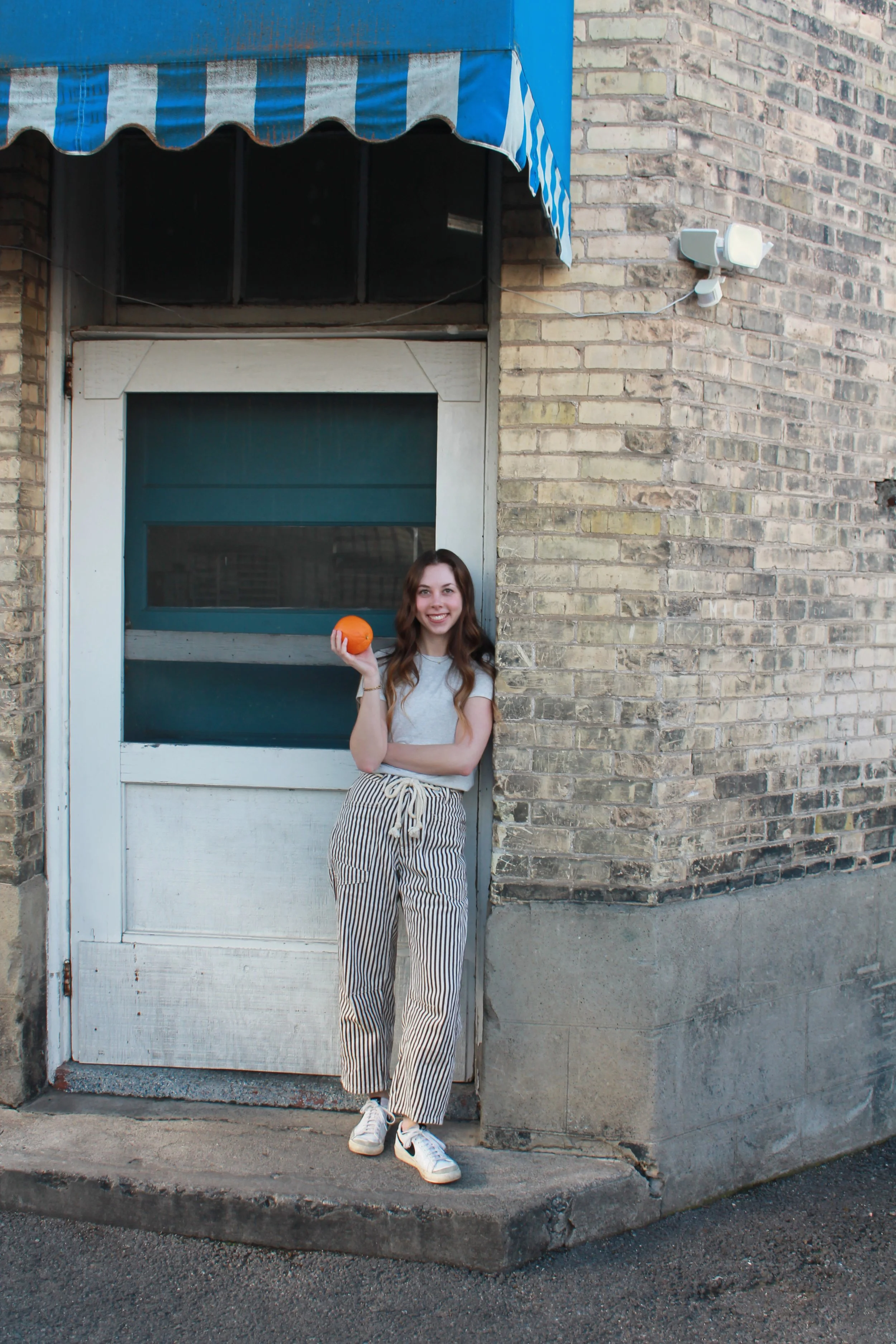 Macy with long brown hair holding an orange fruit while standing in front of a brick building with a blue and white striped awning and an old white door.