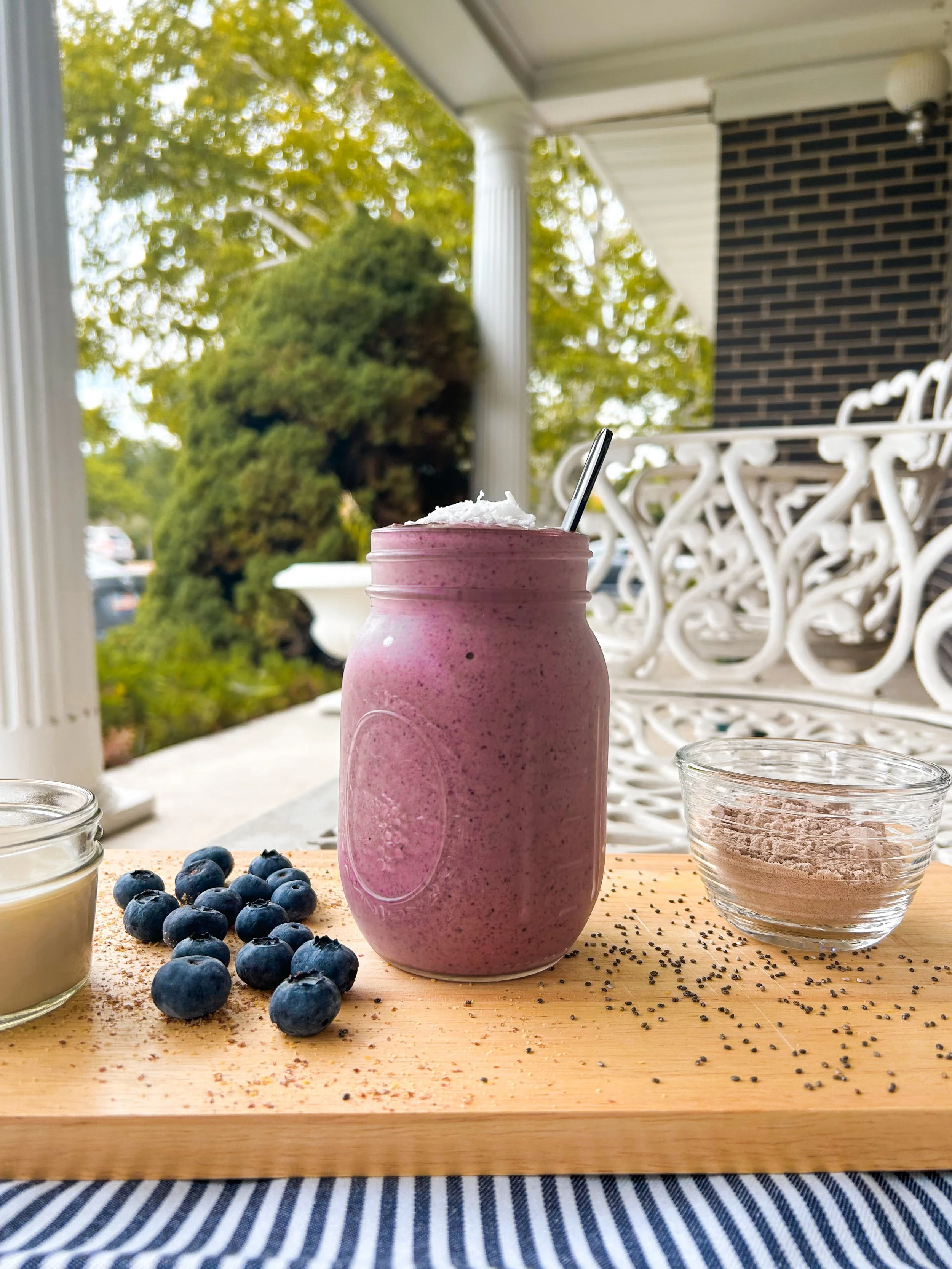 A smoothie in a glass jar on a wooden cutting board, surrounded by fresh blueberries, a small jar of milk, and a glass bowl of protein powder. The background features a porch with white benches and trees with green leaves.