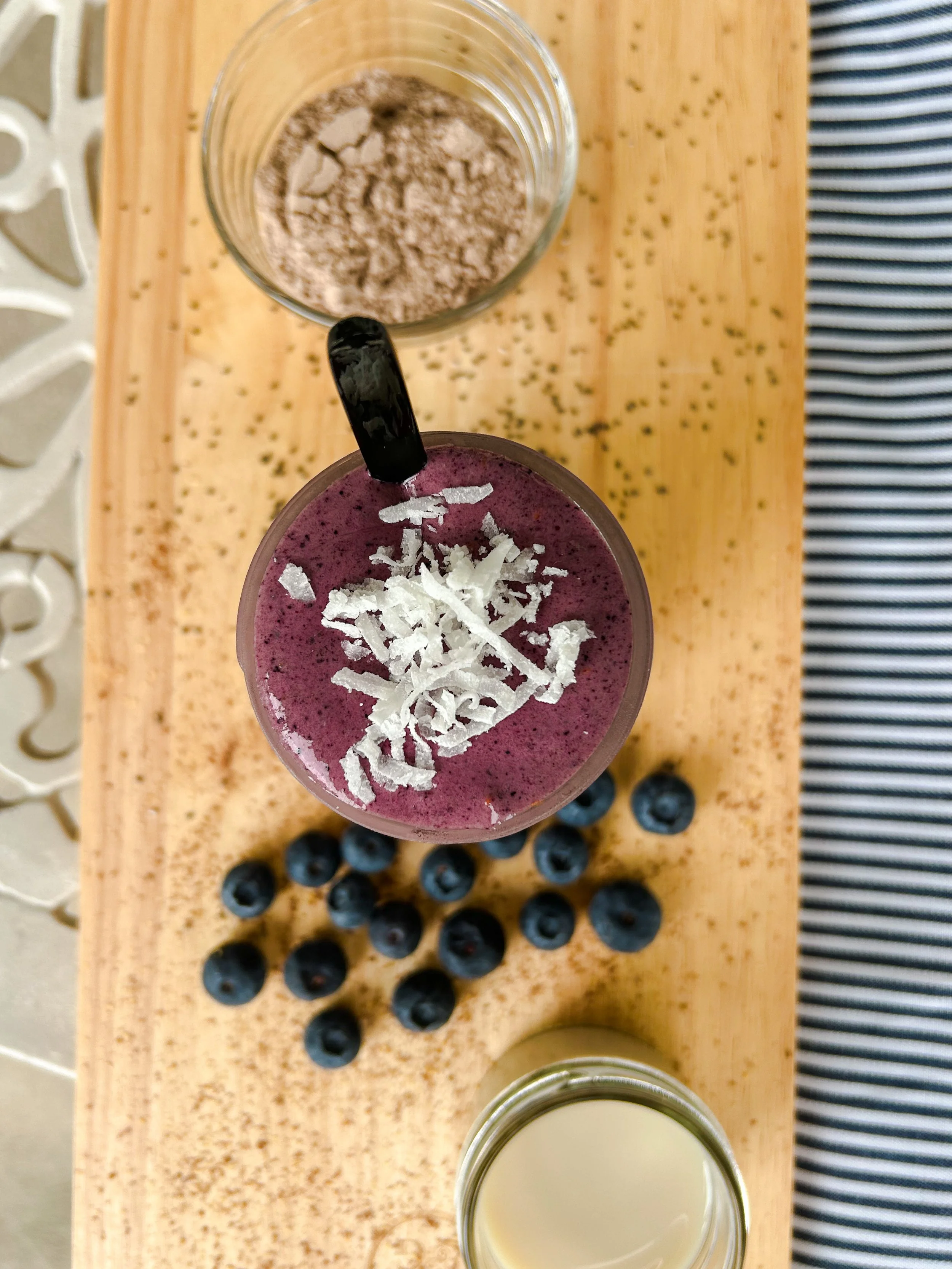 Top view of a smoothie topped with shredded coconut, a jar of milk, a jar of protein powder, and scattered blueberries on a wooden surface.