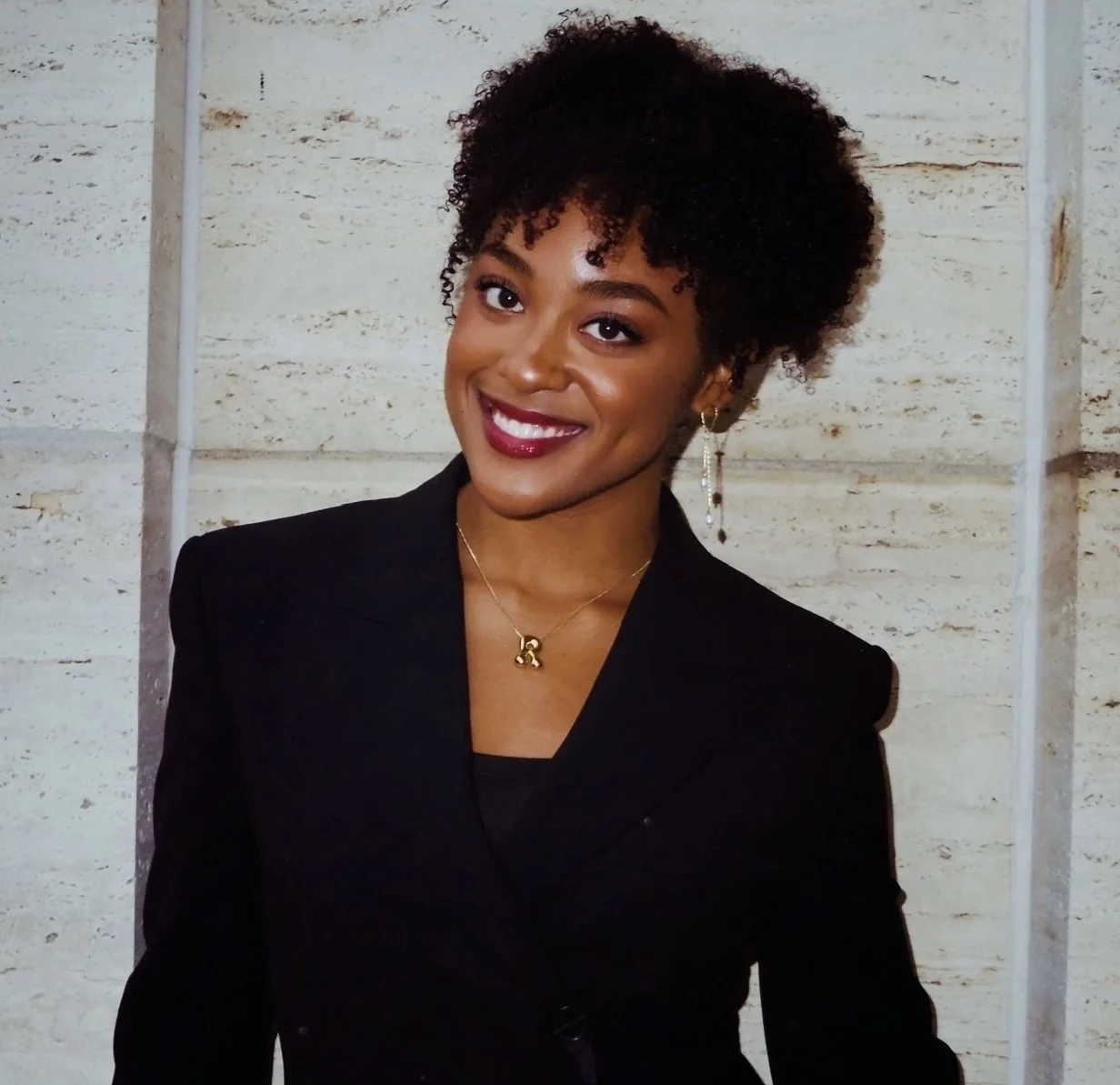 Remidy Destyn Dixon with short, curly black hair, wearing a black blazer, gold necklace, and dangling earrings, standing against a light-colored stone wall.