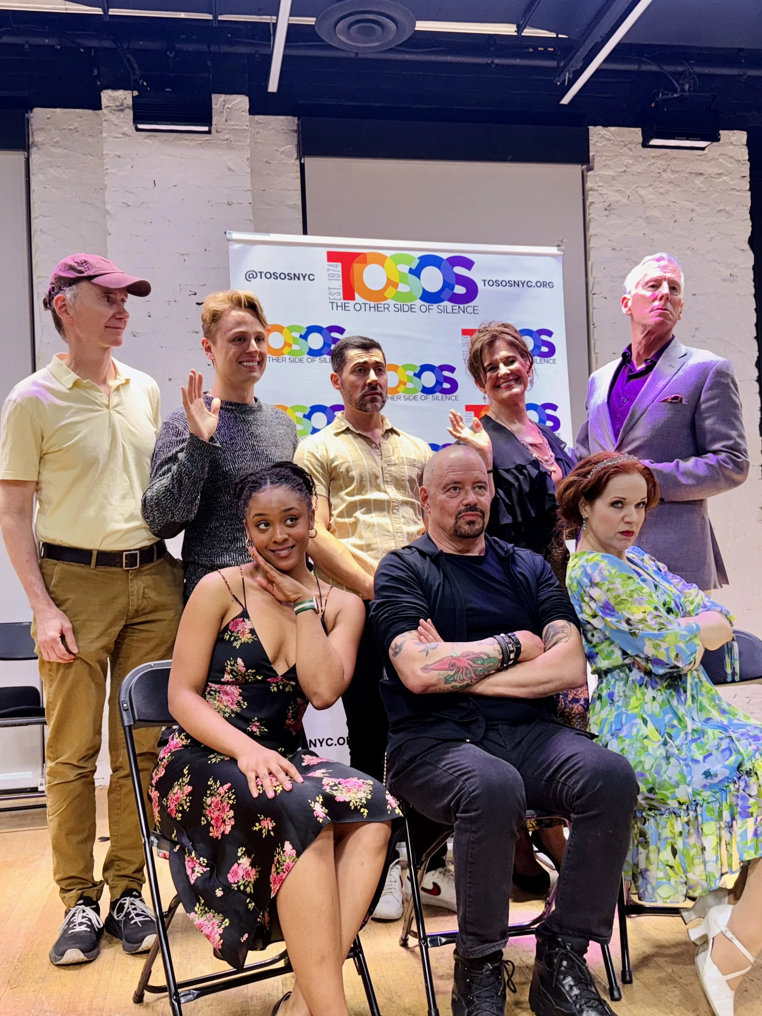 Remidy Destyn Dixon with the cast at TOSOS, an LGBTQ+ organization. Four are seated in front, and five are standing behind. The background includes a white brick wall and a TOSOS banner with rainbow-colored text.