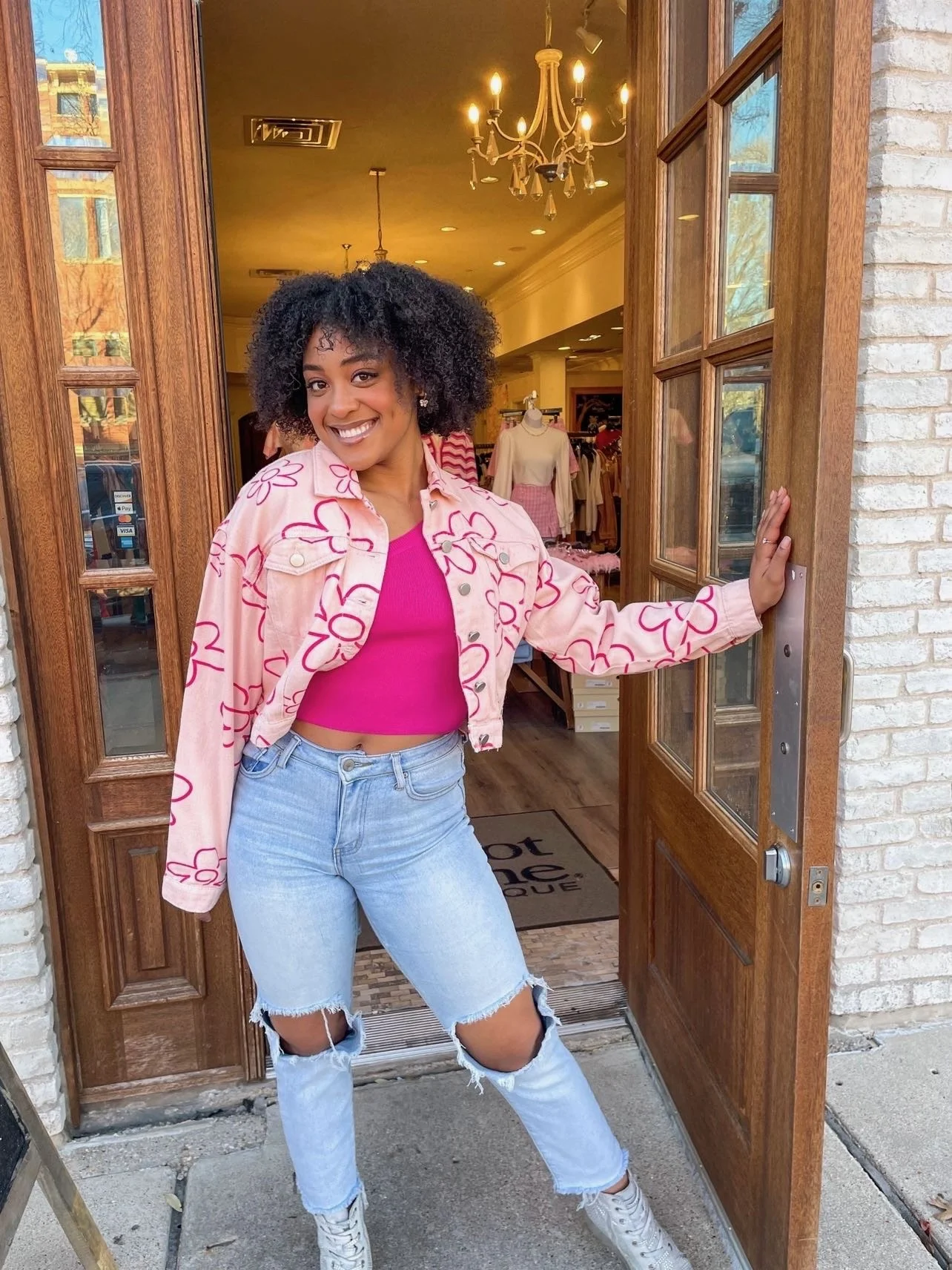Remidy Destyn Dixon with curly hair smiling and standing in a doorway of a clothing store, wearing a pink crop top, a pink jacket with floral patterns, distressed jeans, and white sneakers.