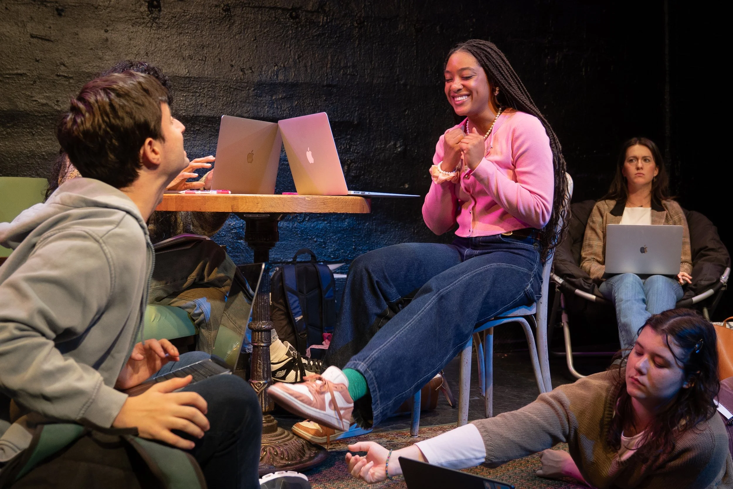 Remidy Destyn Dixon in "In Chief" - A group of young people in a casual setting, with one girl smiling excitedly at a boy sitting on the floor. Some others are working on laptops, and one girl is lying on the floor using her phone.