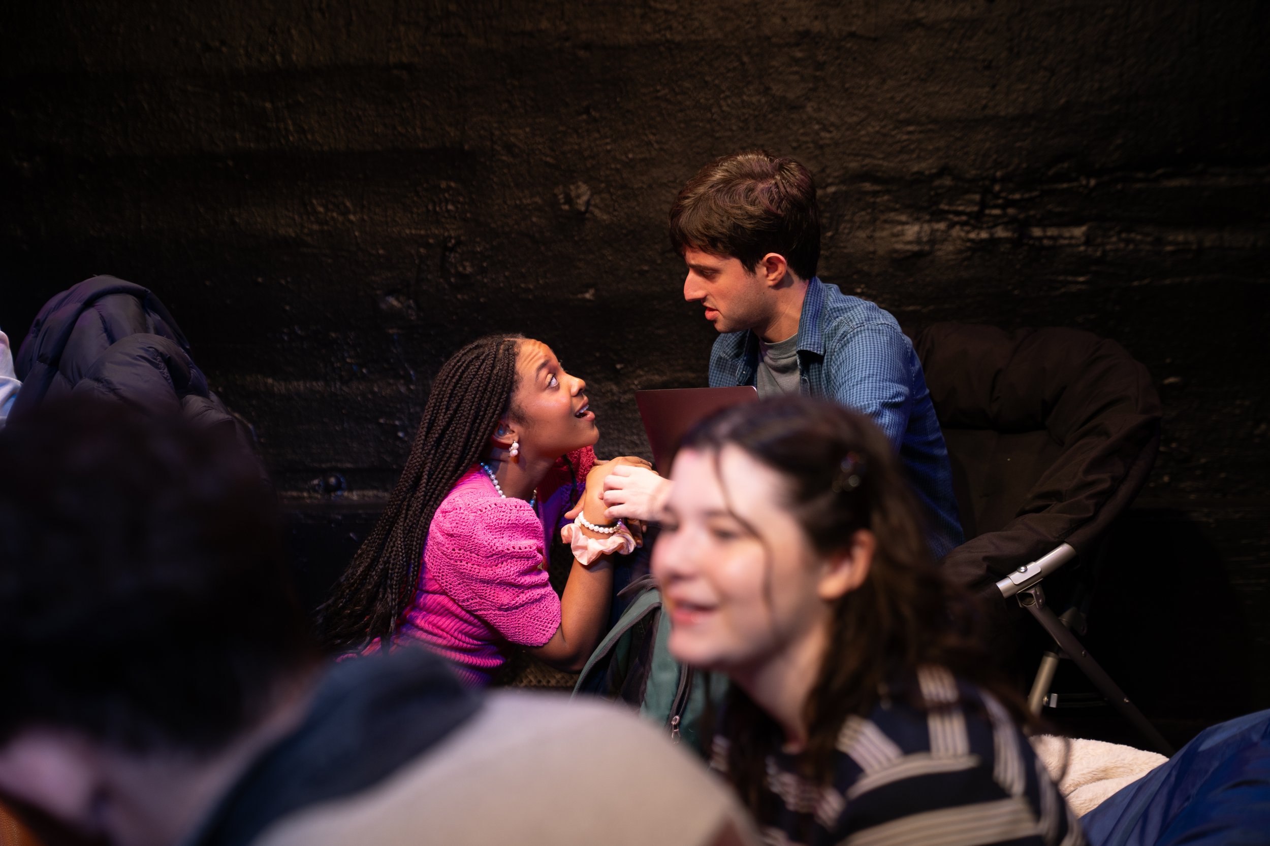 Remidy Destyn Dixon in "In Chief" - A young woman with braided hair wearing a pink sweater is talking to a young man with brown hair wearing a blue shirt, inside a dark room, possibly an underground space.