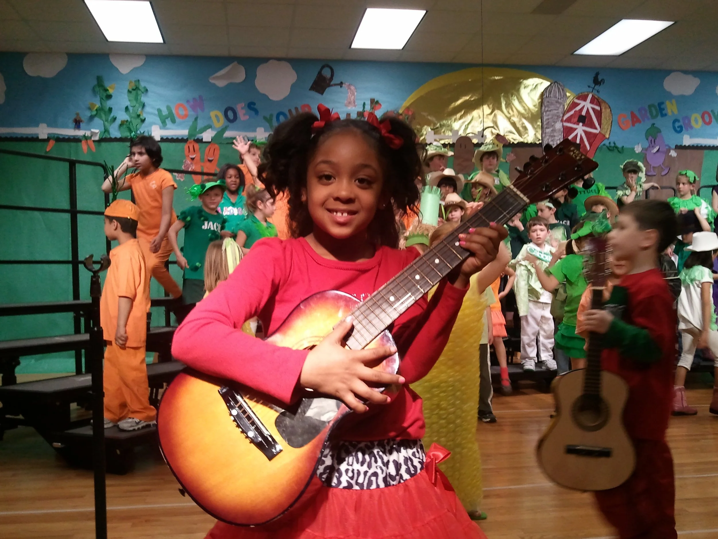 Remidy Destyn Dixon  as a Young girl in a pink shirt and animal-print skirt holding a guitar, smiling at camera during a school play with children in colorful costumes on stage in the background.