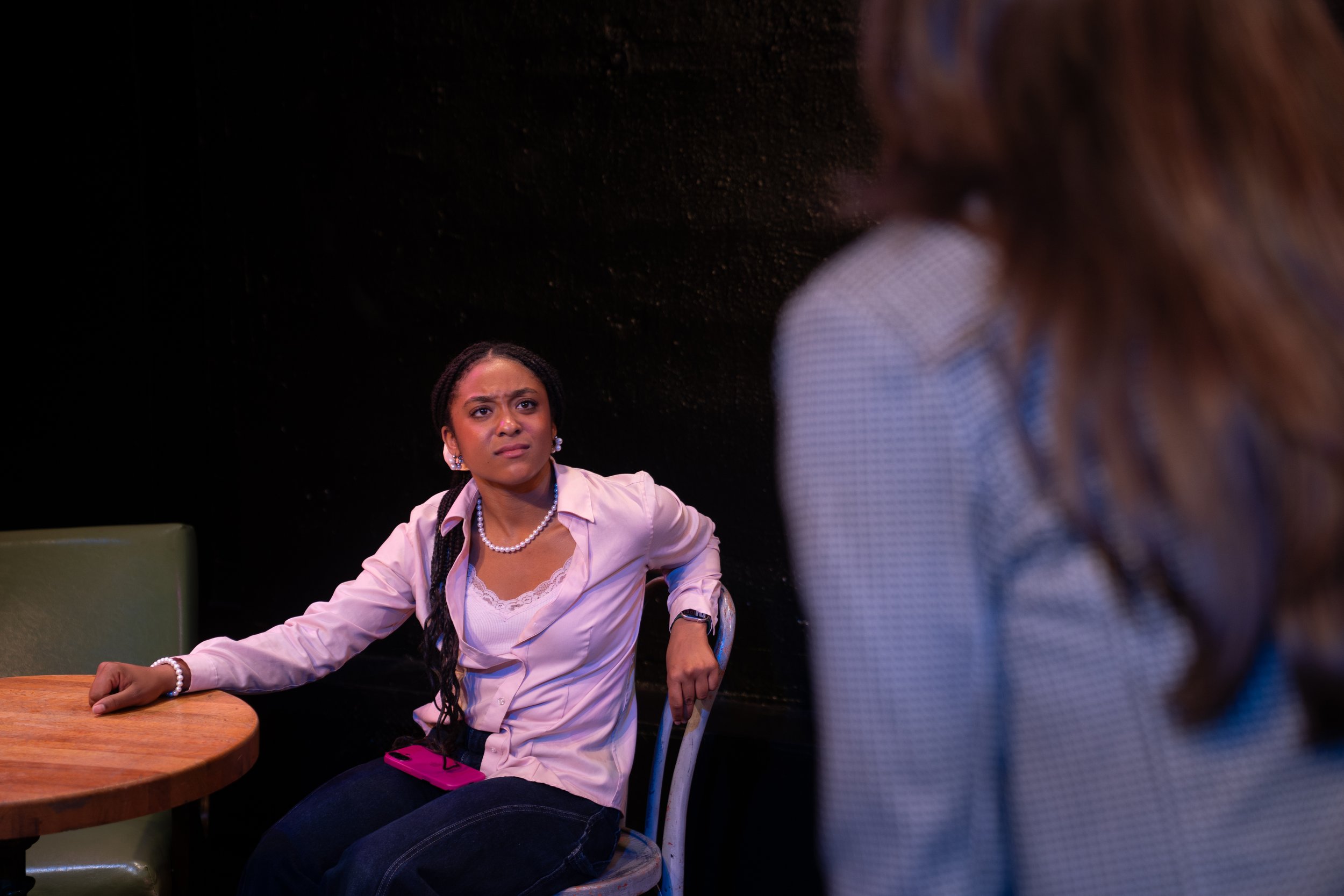 Remidy Destyn Dixon in "In Chief" - A woman with braided hair, wearing a white shirt and pearl jewelry, sitting at a wooden table with a serious or confused expression, having a conversation.