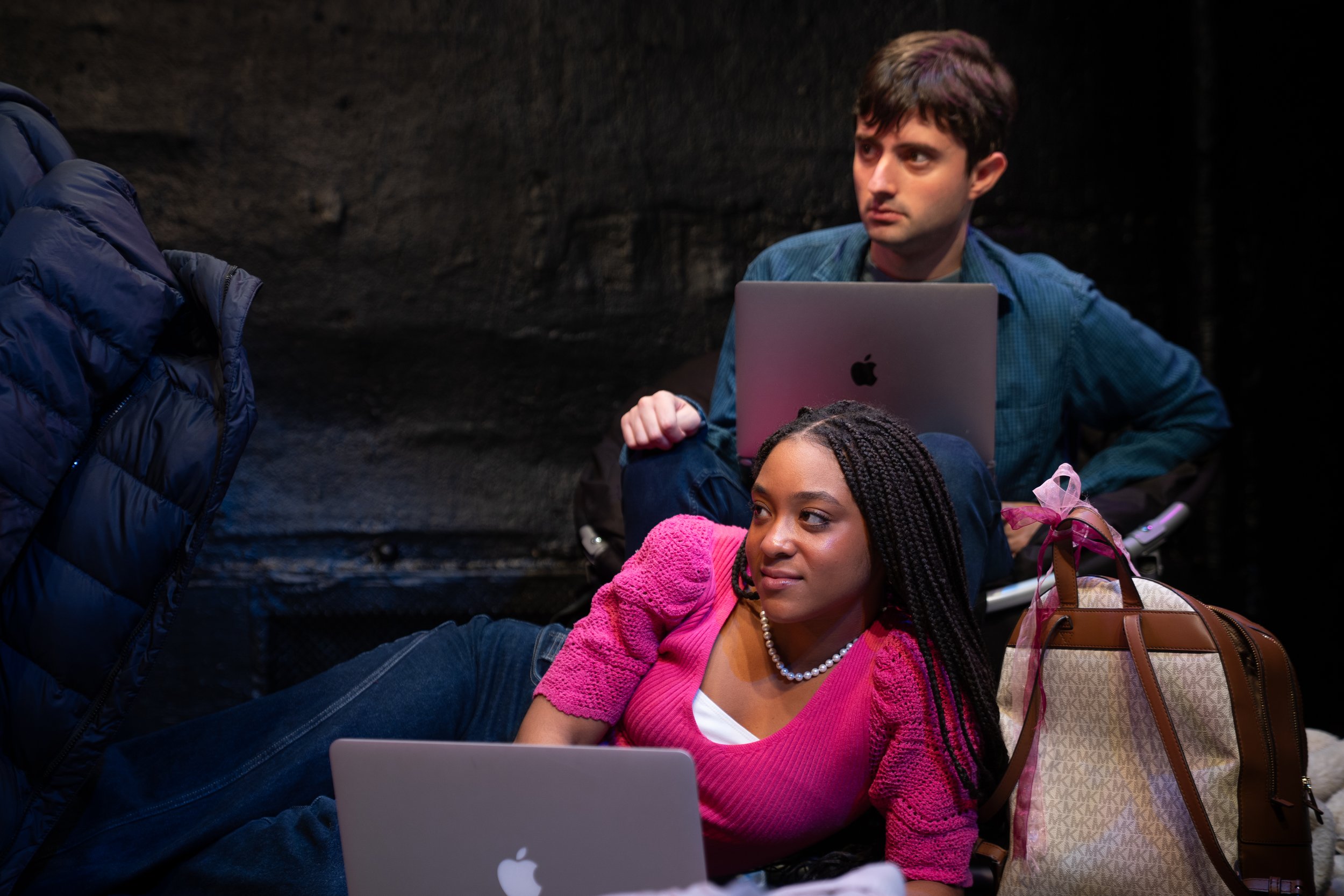 Remidy Destyn Dixon in "In Chief" - Two young people relaxing with laptops in a dark indoor setting. The woman is lying down, wearing a pink sweater and pearl necklace, with a branded bag nearby. 