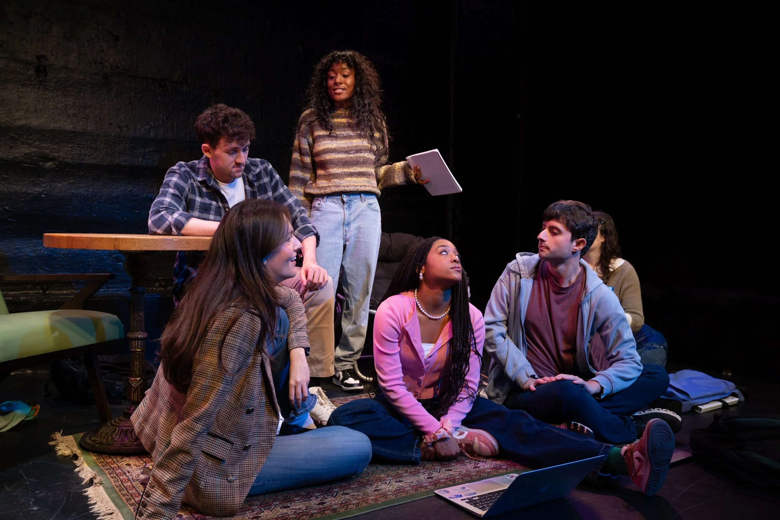 Remidy Destyn Dixon in "In Chief" Six young people gathered in a stage acting scene, some sitting on the floor, one standing, one at a table, engaged in conversation, with a laptop and backpack nearby.