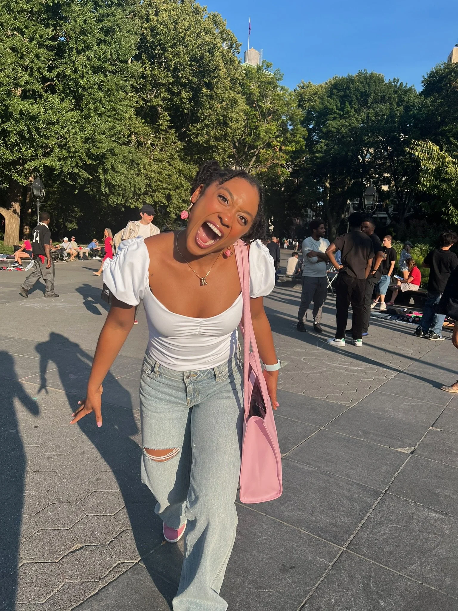 Remidy Destyn Dixon with short dreadlocks and wearing a white top, ripped jeans, and pink sneakers, smiling and leaning forward in an outdoor city park setting with numerous people, trees, and a tall building in the background.