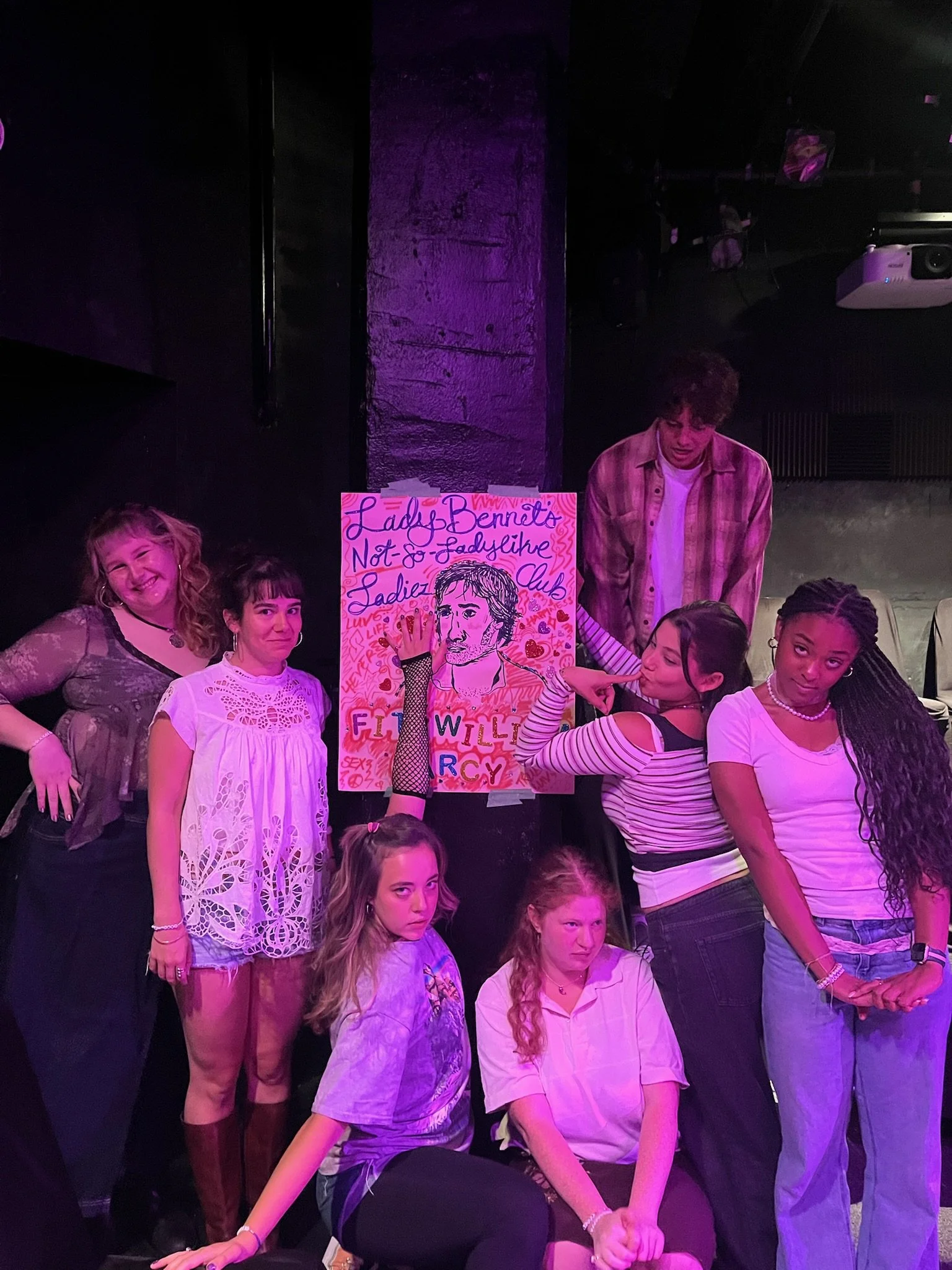 Remidy Destyn Dixon with the cast of Ladiez of P&P, written by Laini Sharifi standing and sitting around a colorful poster with a drawing and handwritten text, in a dimly lit room with purple lighting.