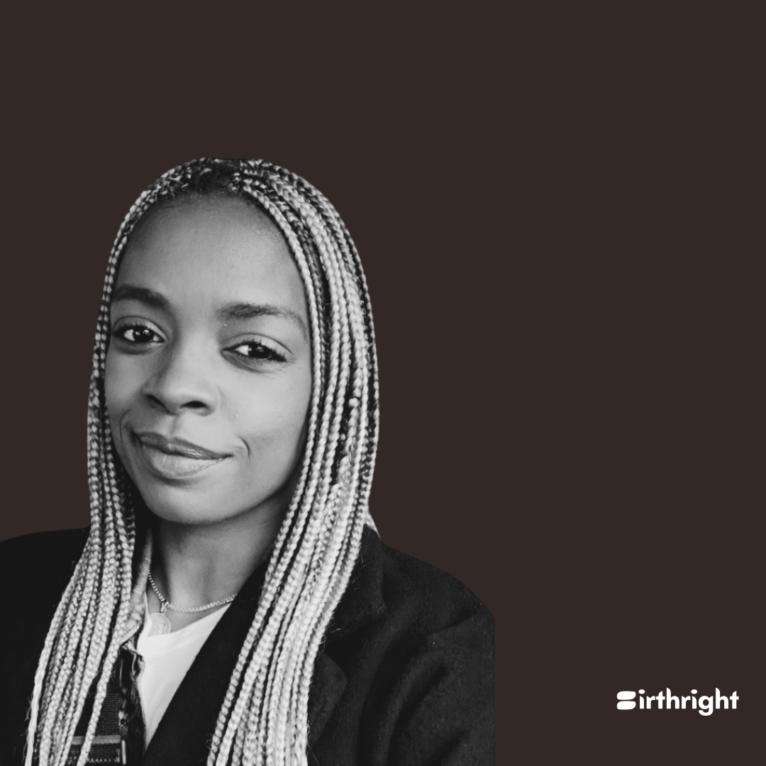 Black and white photo of a woman wearing a wide-brim hat, a necklace, and a dark jacket, with braided hair, smiling slightly, against a dark background.