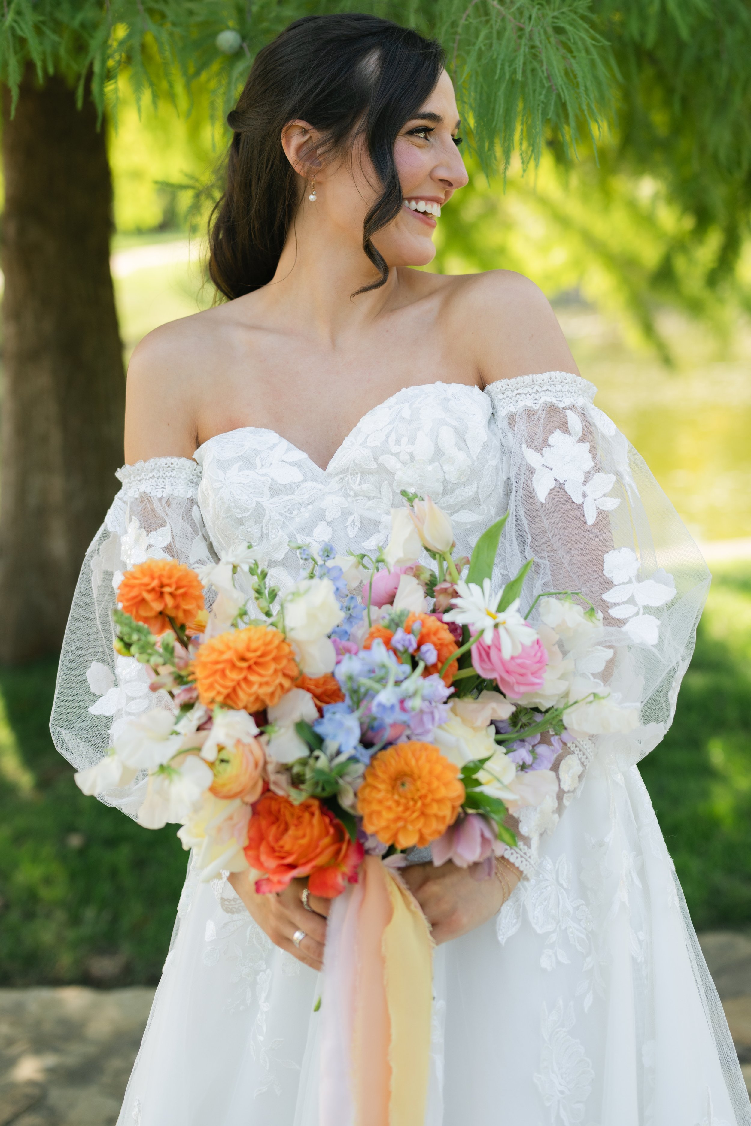 A woman in a white wedding dress holding a colorful bouquet of flowers outdoors with green trees and grass in the background.