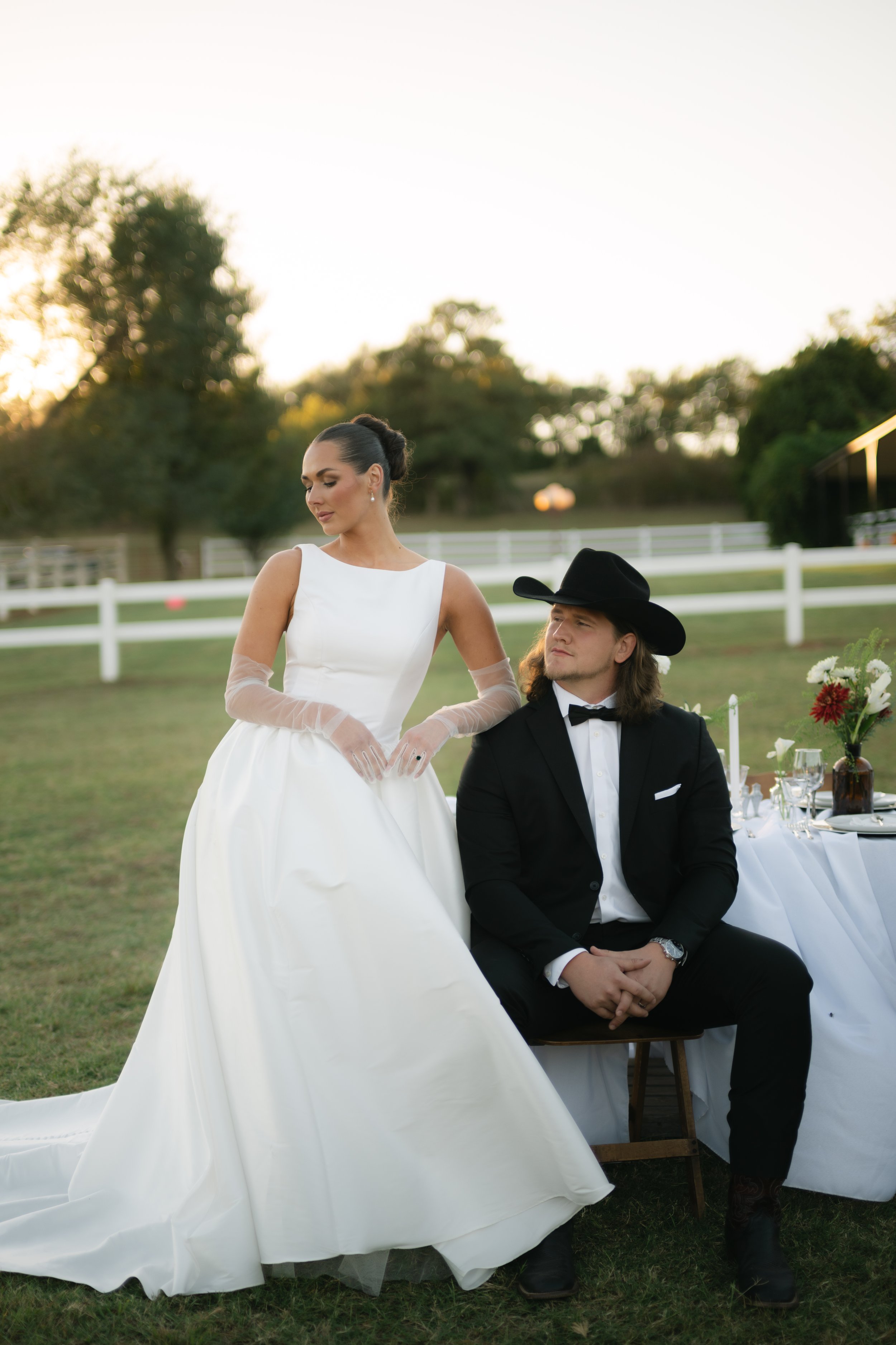 A bride in a white gown with sheer sleeves and a groom in a black tuxedo and cowboy hat sit outdoors near a table with flowers and glassware, during sunset.