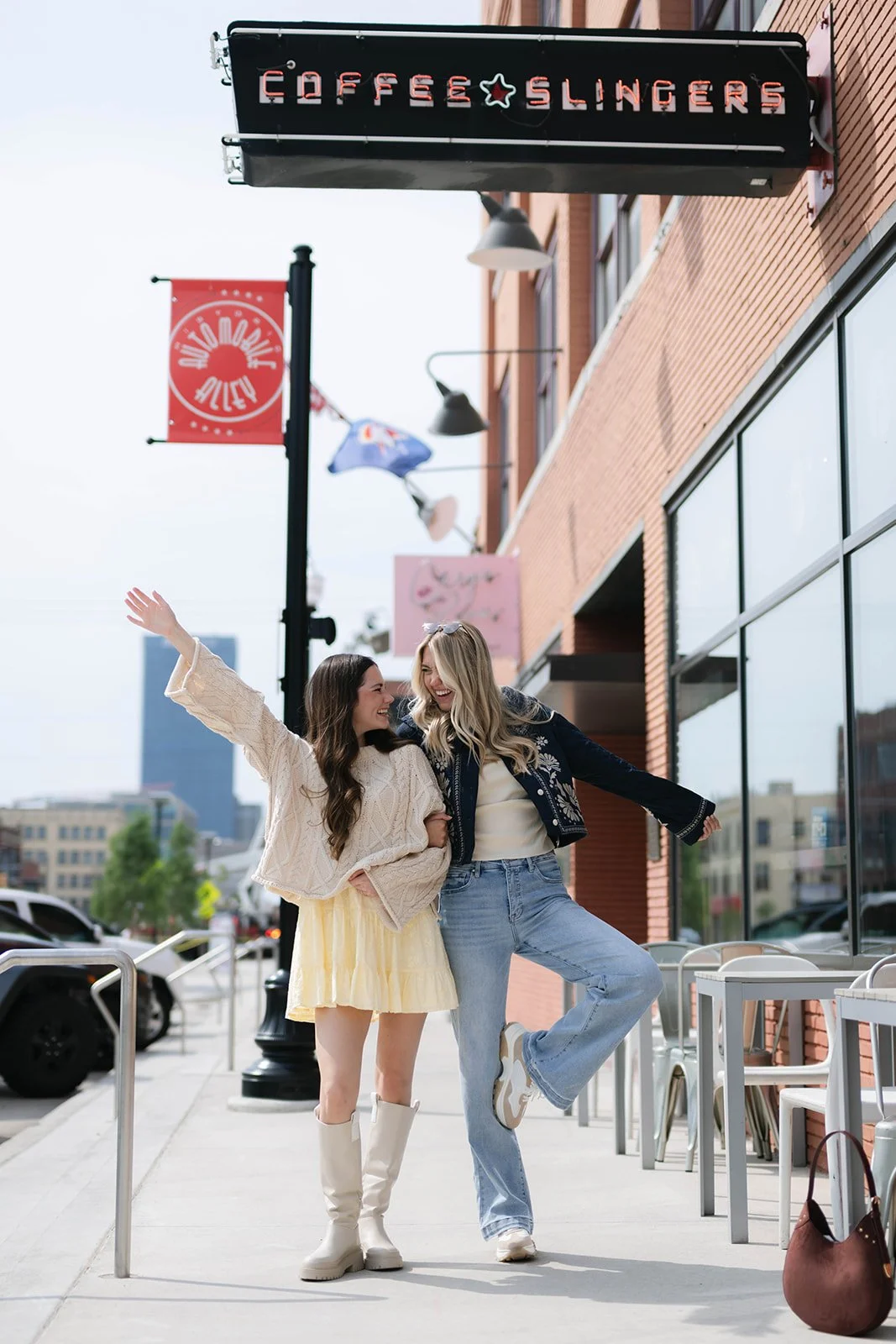 Two young women smiling and enjoying themselves on a city sidewalk in front of a cafe with a sign that reads 'Coffee Slangers.'