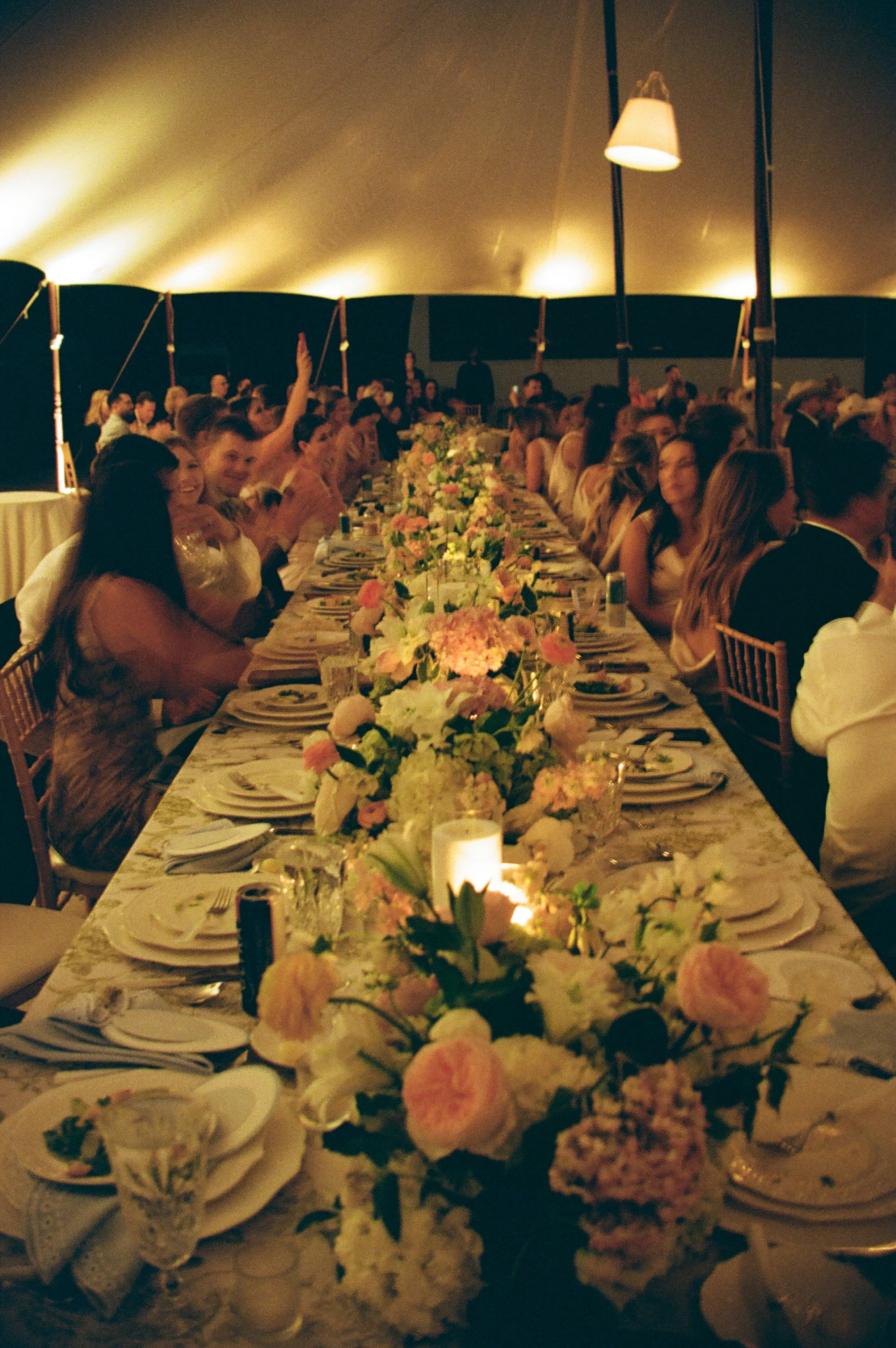 A long banquet table decorated with pink and white flowers, lit by candles, with people seated on both sides, inside a large tent at an event or celebration.