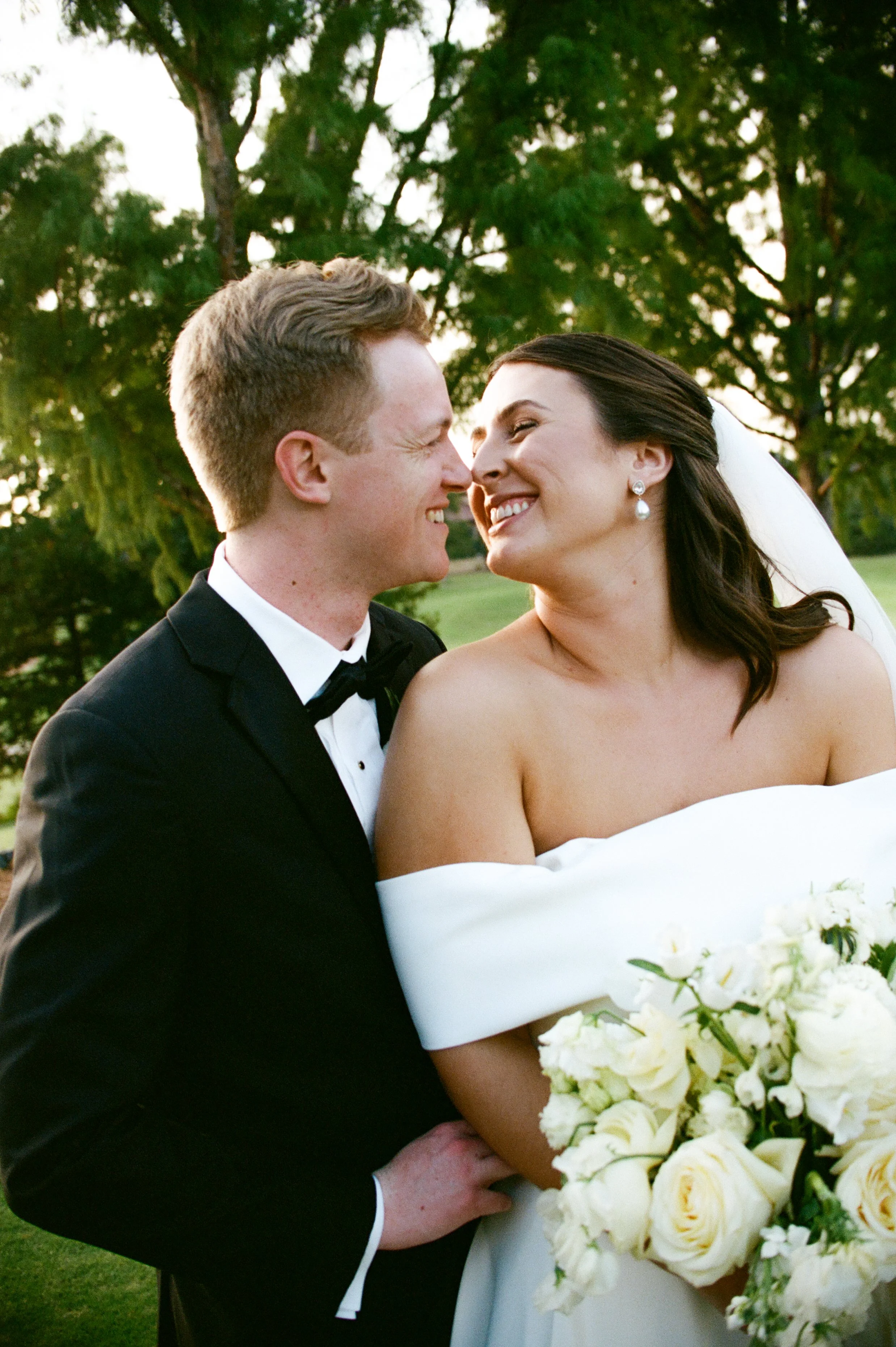 A newlywed couple smiling and touching noses outdoors, with trees in the background. The groom is in a black tuxedo and bow tie, the bride is in a strapless white wedding dress, holding a bouquet of white roses.