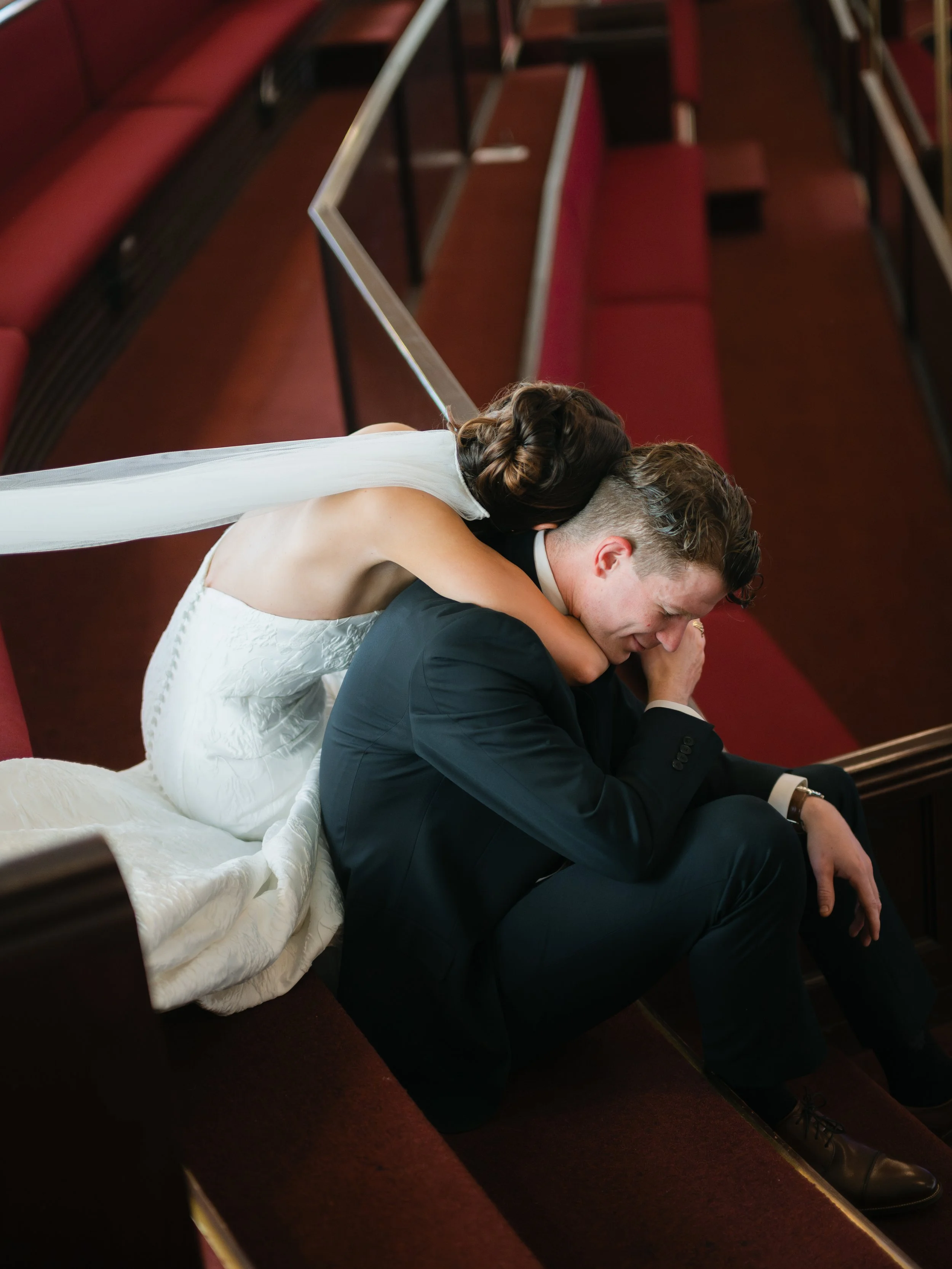 A bride and groom sitting on the steps of a church, with the bride hugging the groom from behind as he sits with eyes closed and hands clasped near his face.
