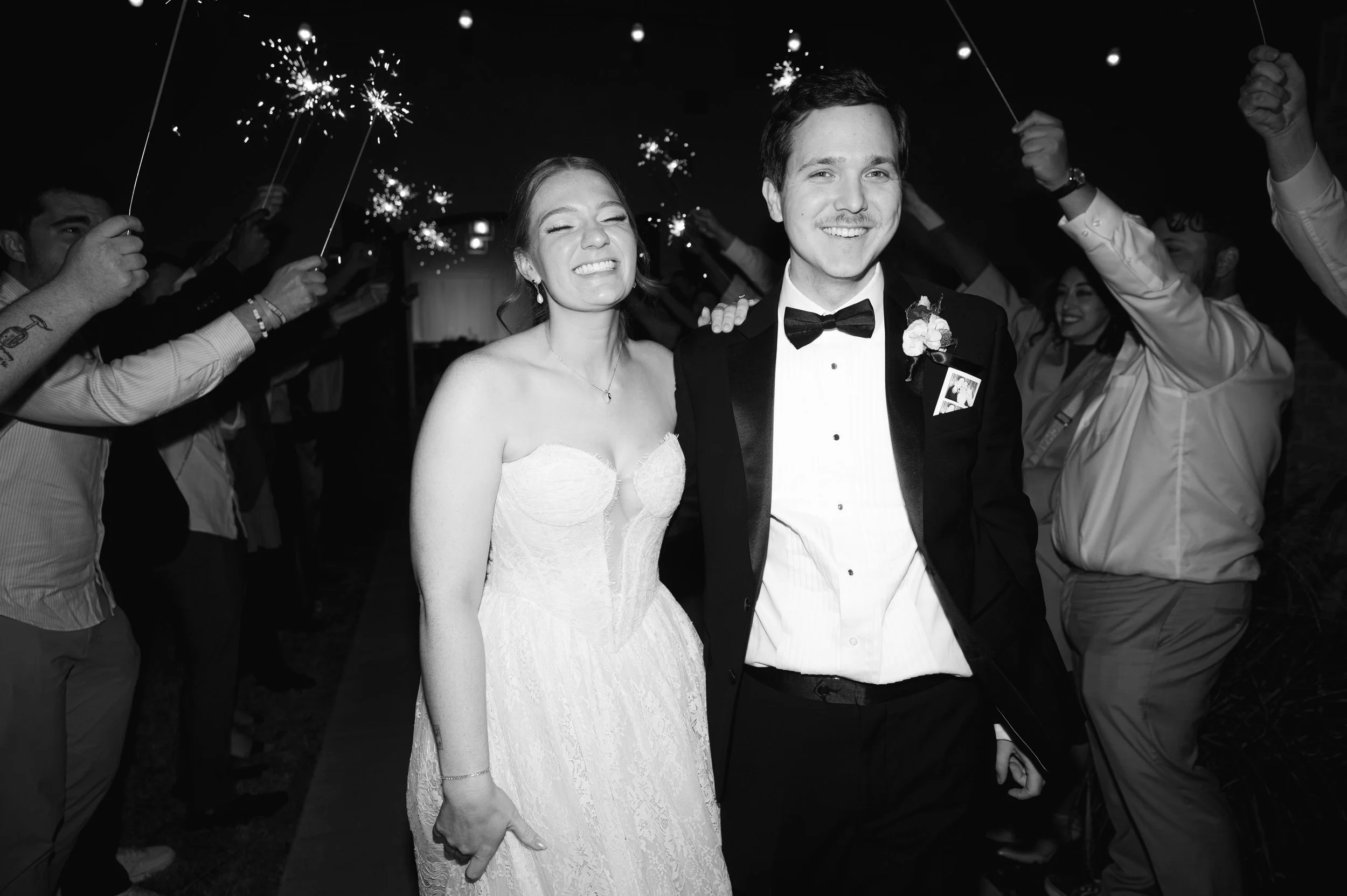 Black and white photo of a newlywed couple smiling as they walk through a crowd holding sparklers during their wedding celebration.