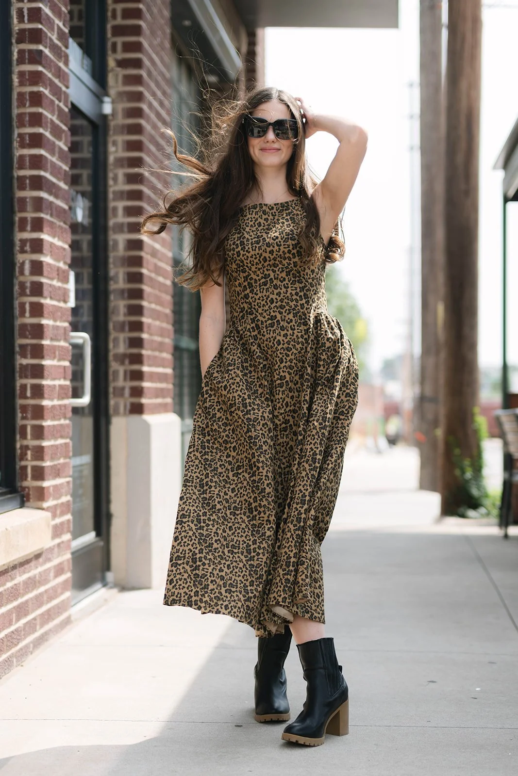 A woman with long wavy brown hair, wearing black sunglasses, a leopard print sleeveless dress, and black platform boots, standing outdoors on a sidewalk next to a brick building with large windows and wooden support beams.