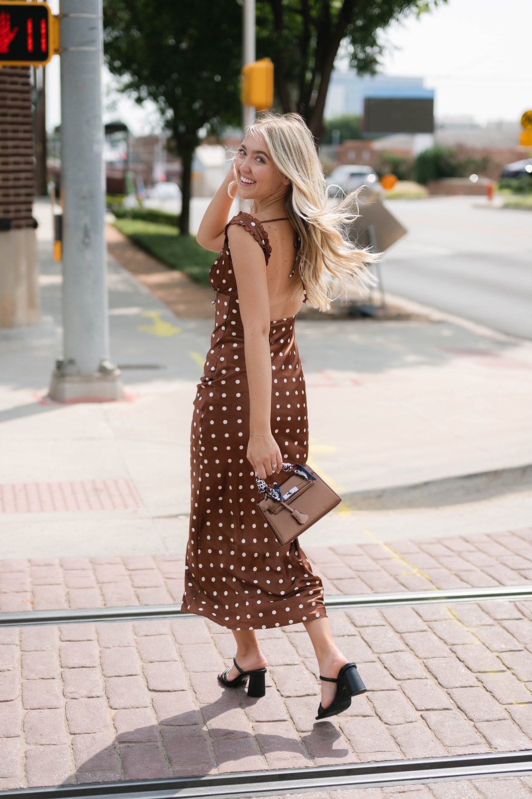 A woman in a brown polka dot dress walking on a city sidewalk, smiling and looking back. She has long blonde hair and is holding a small brown handbag.