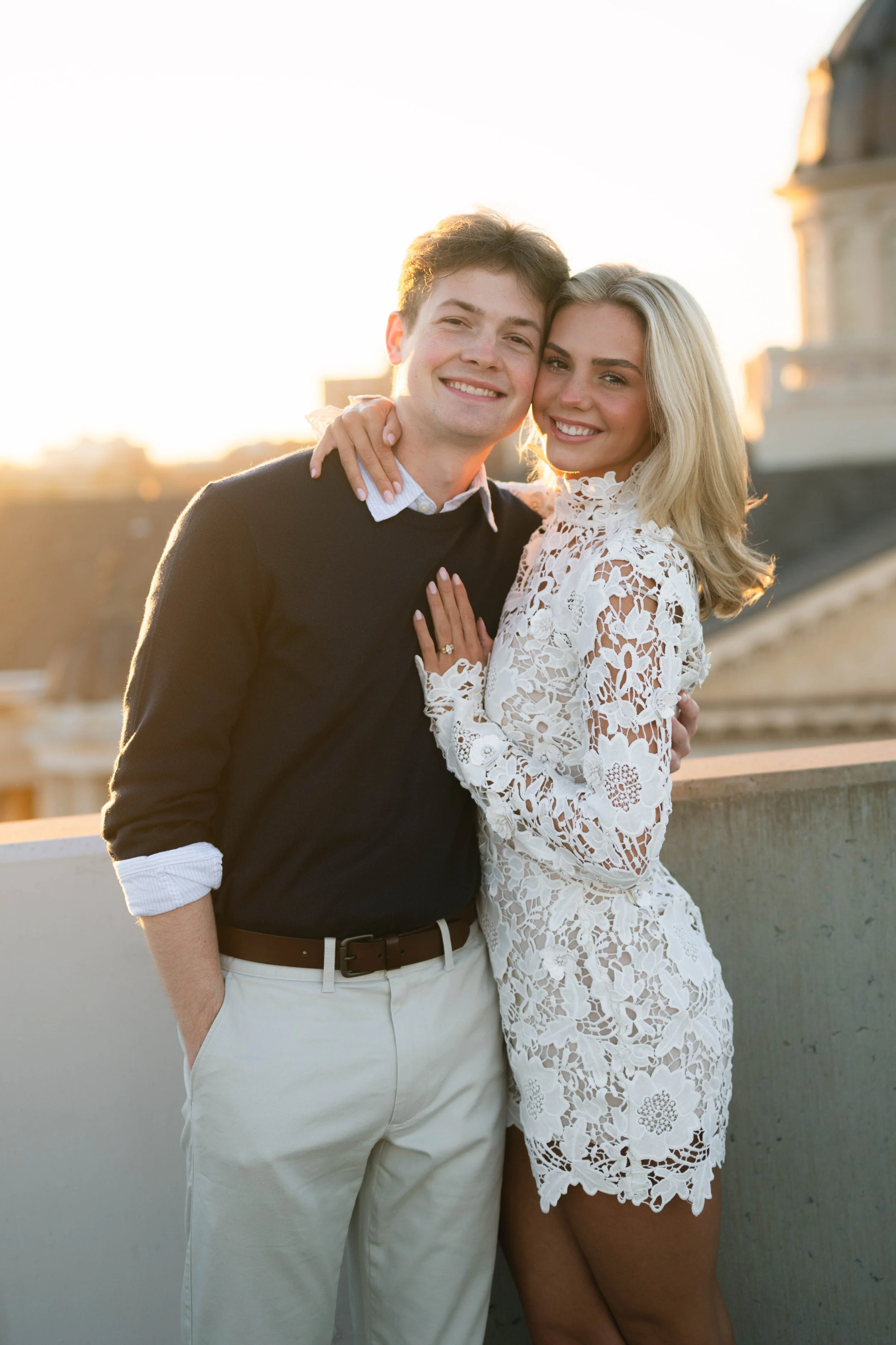 A happy couple smiling and posing together outdoors at sunset, with a cityscape background.