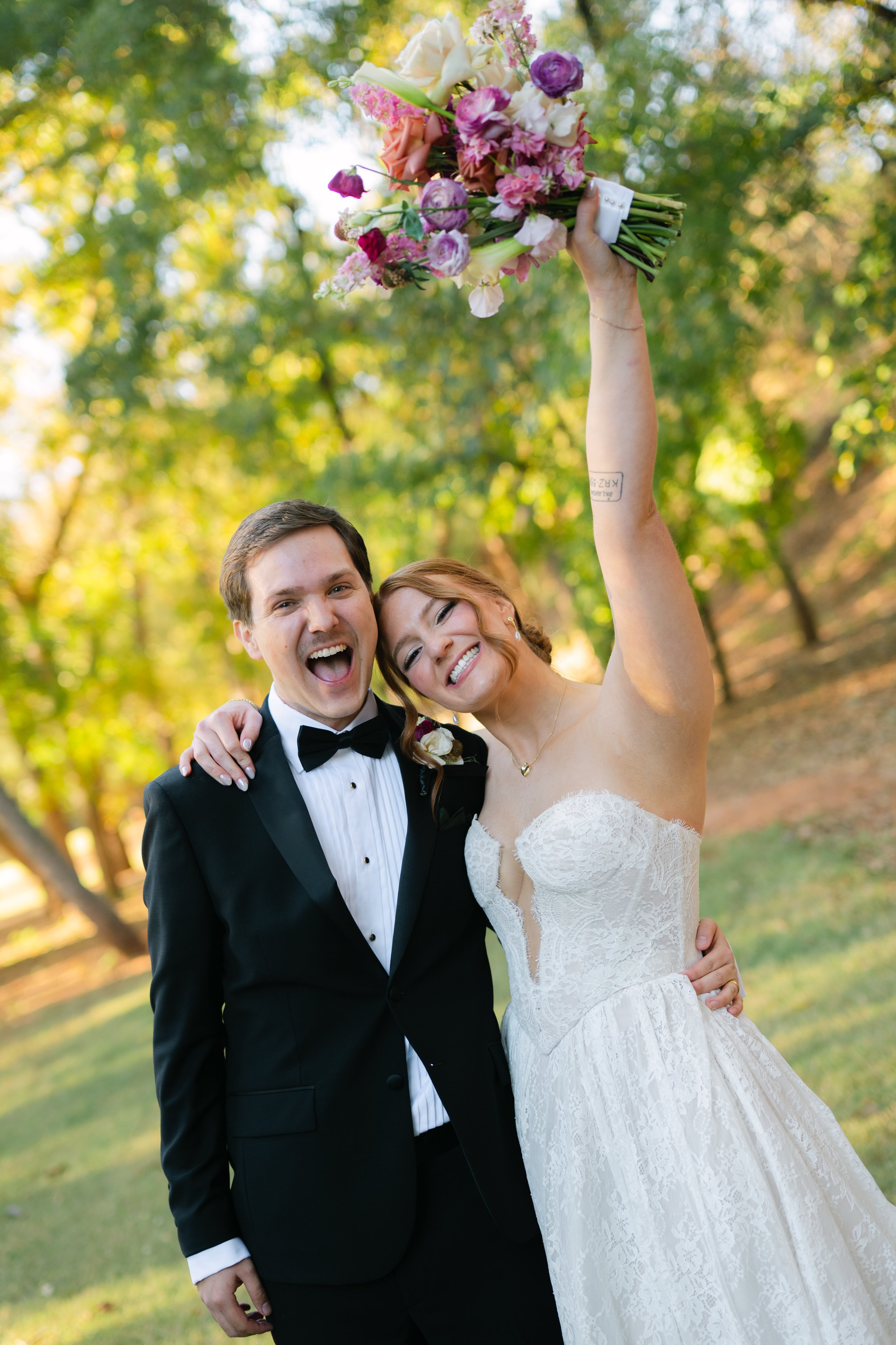Happy bride and groom smiling outdoors, the bride holding a colorful bouquet in the air.