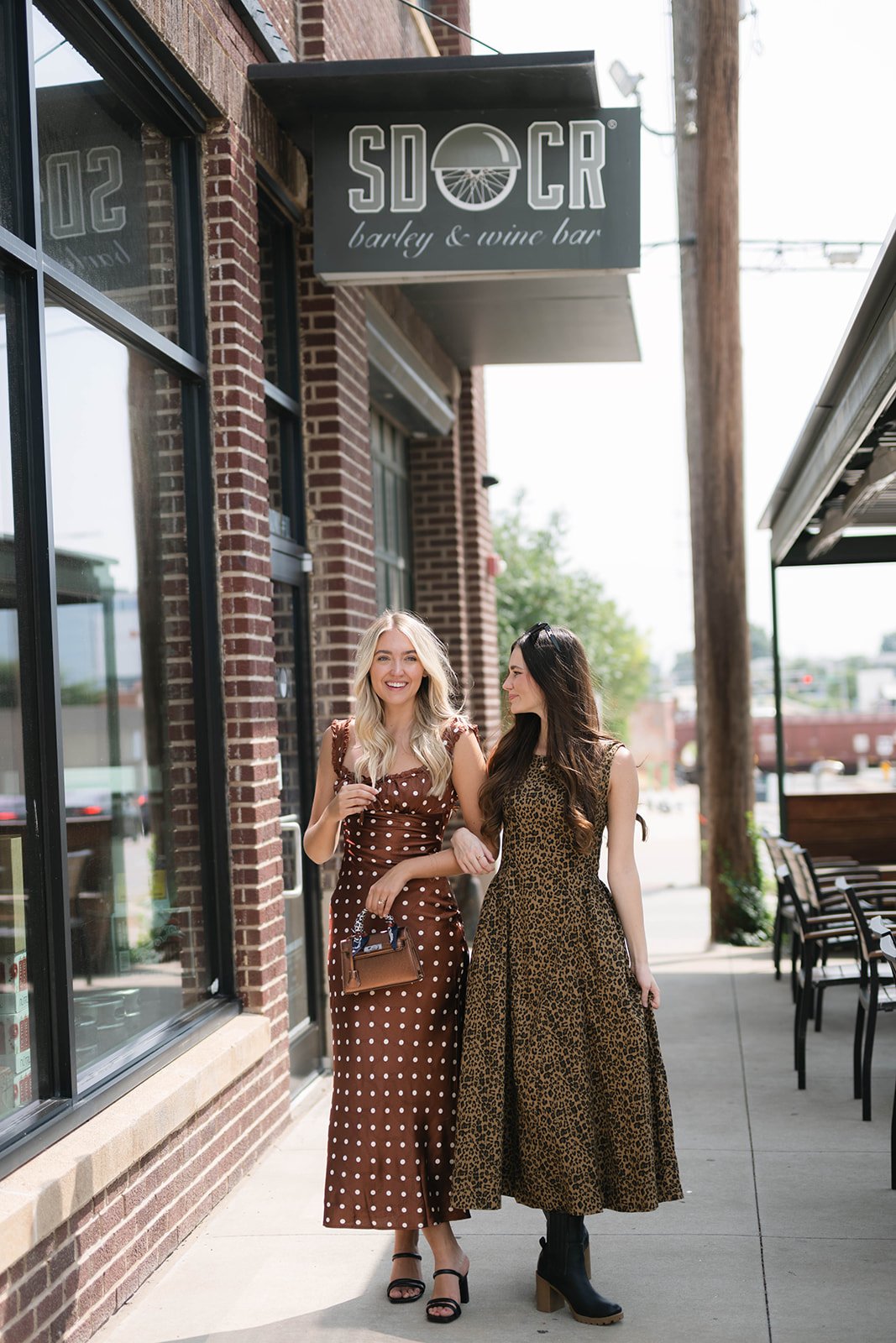 Two women walking arm-in-arm outside of a bar called SD OCR, which offers barley and wine. They are dressed stylishly, one in a brown polka dot dress and the other in a leopard print dress, smiling and enjoying each other's company.