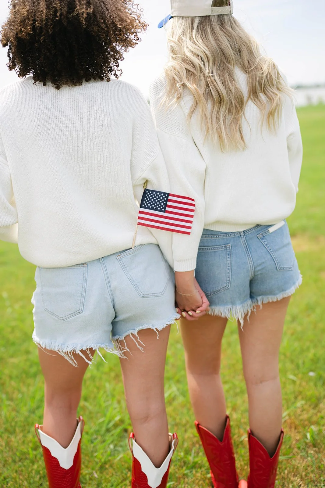 Two women dressed in white sweaters and denim shorts stand outdoors on green grass, holding hands. One woman has an American flag patch on her sleeve. They are wearing cowboy boots.