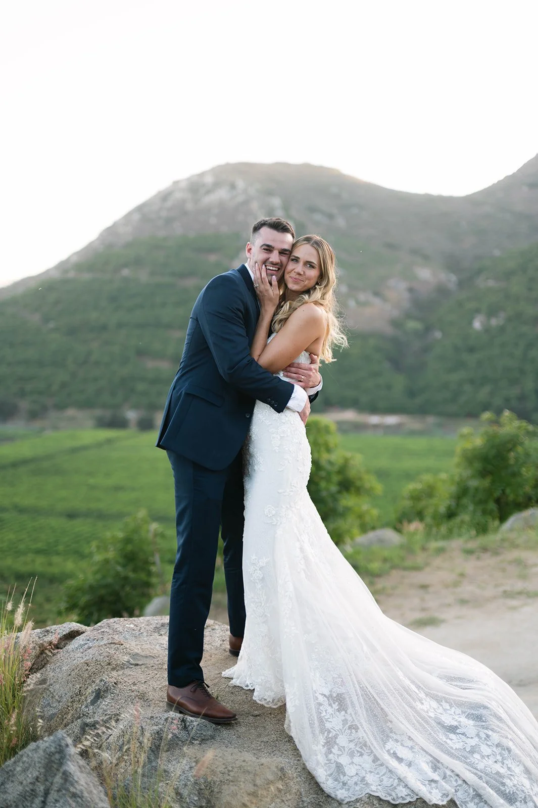 A newlywed couple in wedding attire sharing a joyful embrace outdoors with mountains and greenery in the background.