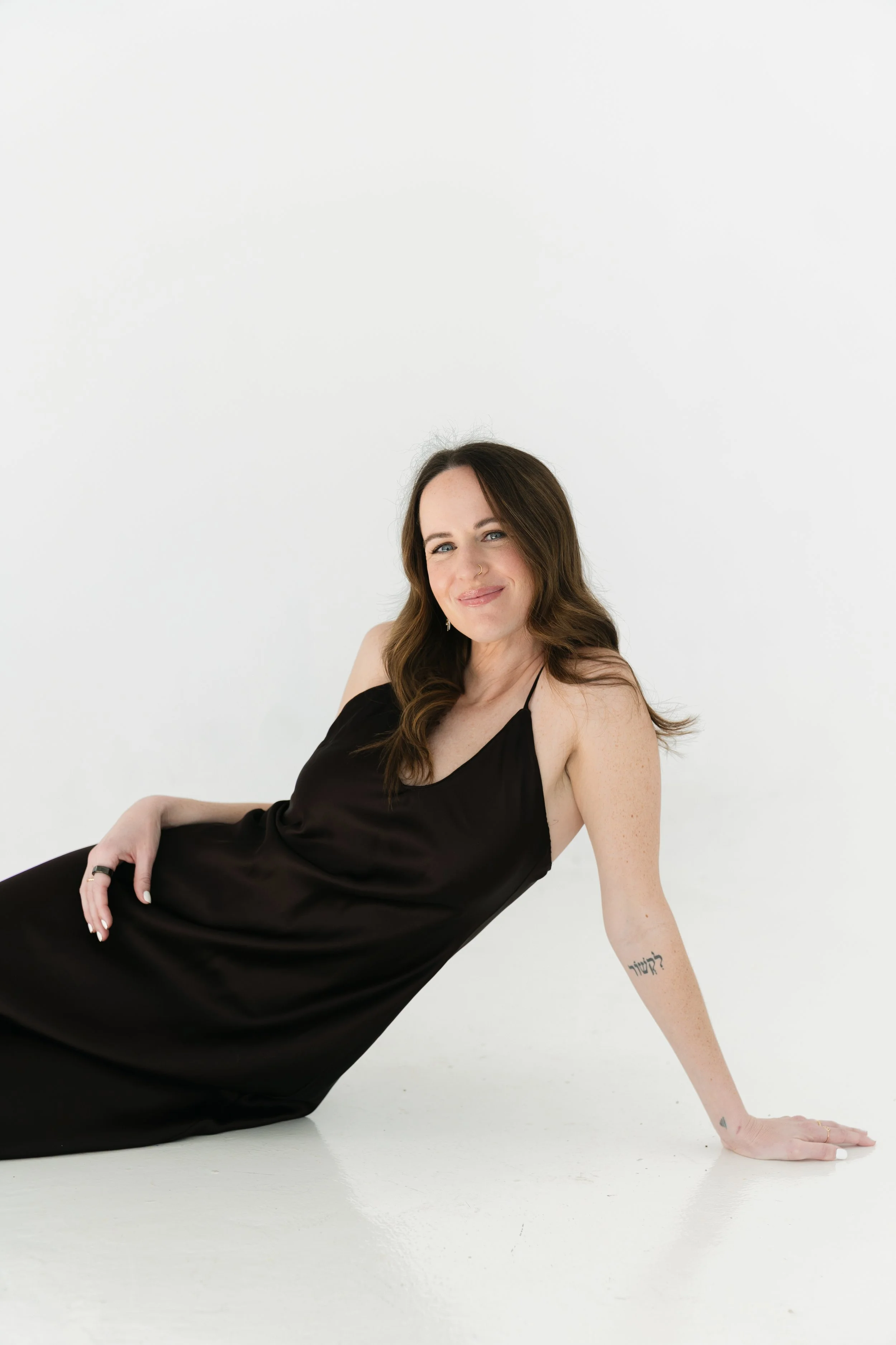 A woman with long brown hair wearing a black dress, sitting on the floor and smiling at the camera against a plain white background.